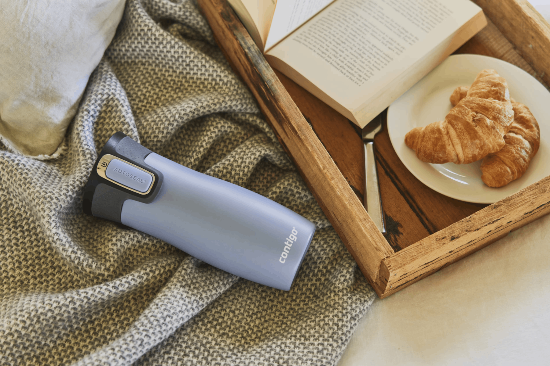 Taza de viaje Contigo AUTOSEAL azul sobre una bandeja con un libro y cruasanes. Manta gris de fondo, luz suave.