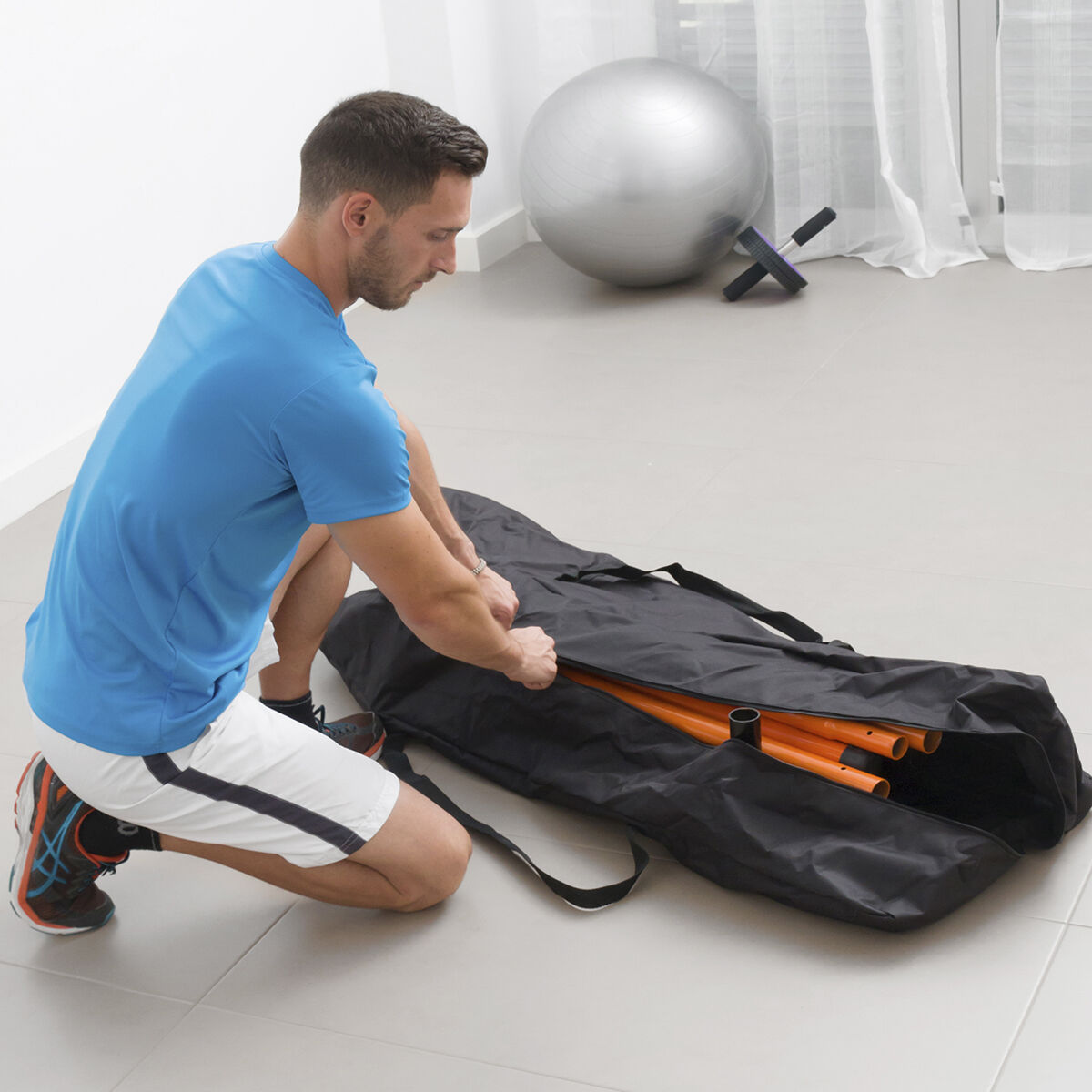 Un hombre arrodillado, abriendo una bolsa negra. Se ven postes naranjas dentro, y una bola de fitness detrás.