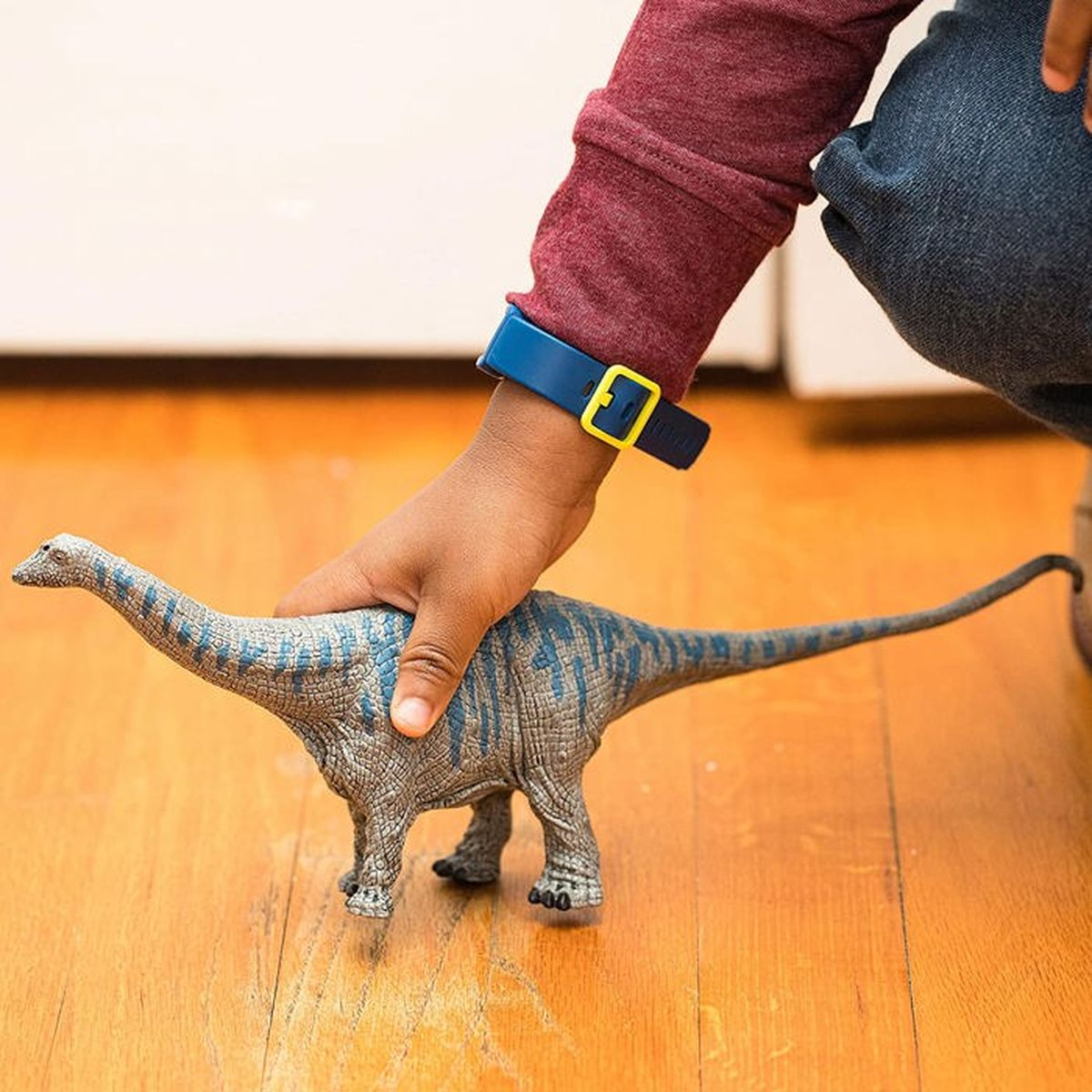 La mano di un bambino tiene un giocattolo a forma di dinosauro su un pavimento di legno. Il bambino indossa un orologio blu.