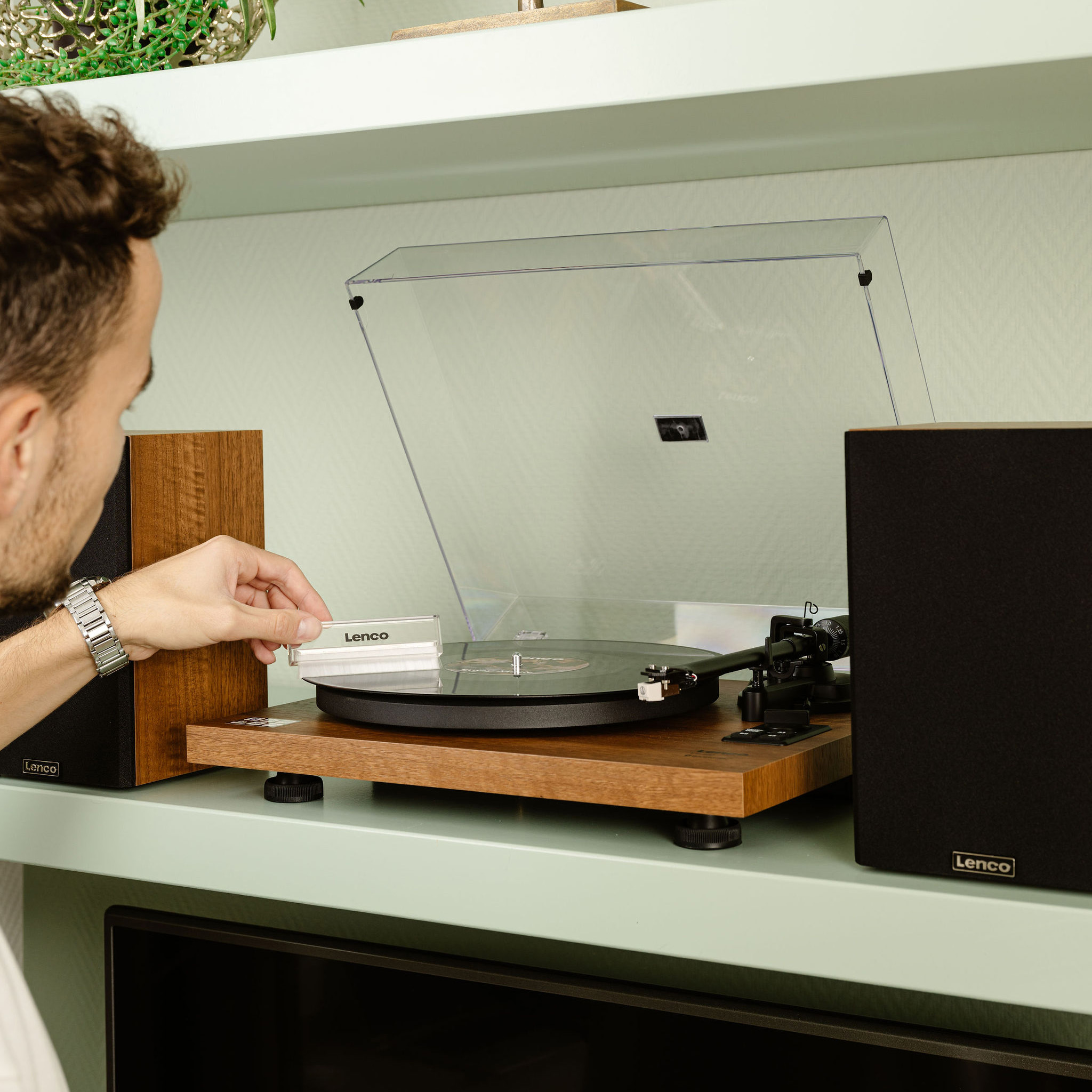 Man using a Lenco turntable, with speakers, on a shelf, in a room setting. 