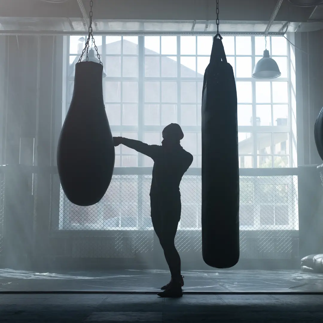 Silhouette einer Person beim Boxen im Fitnessstudio. Zwei hängende Boxsäcke, großes Fenster im Hintergrund.