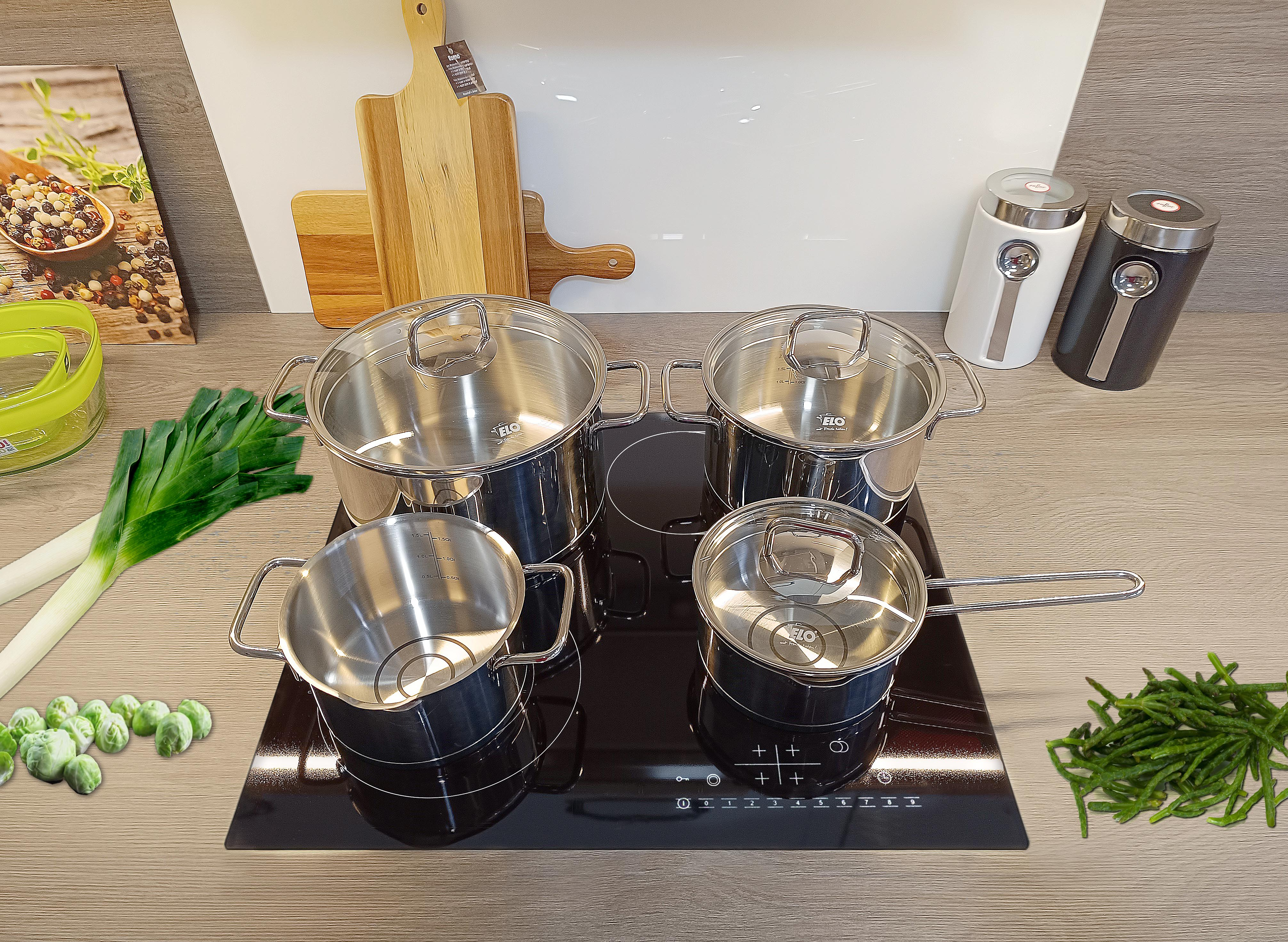 Vier Edelstahltöpfe mit Deckeln auf einem schwarzen Kochfeld, mit Schneidebrettern und Zutaten.