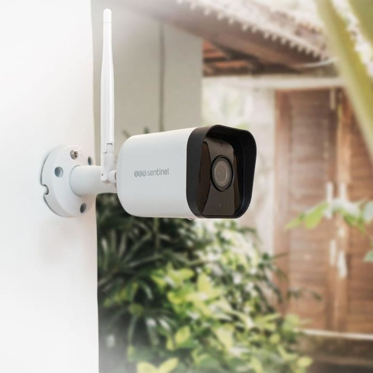 Telecamera di sicurezza bianca montata su un muro. Ha un obiettivo nero e una piccola antenna. Fogliame verde sullo sfondo.