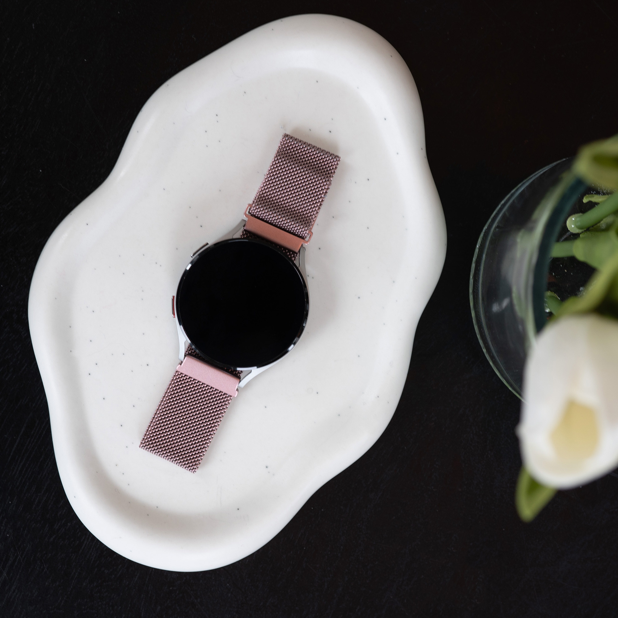 Eine Uhr mit pinkfarbenem Armband auf einem weißen, organisch geformten Teller, auf schwarzem Untergrund.