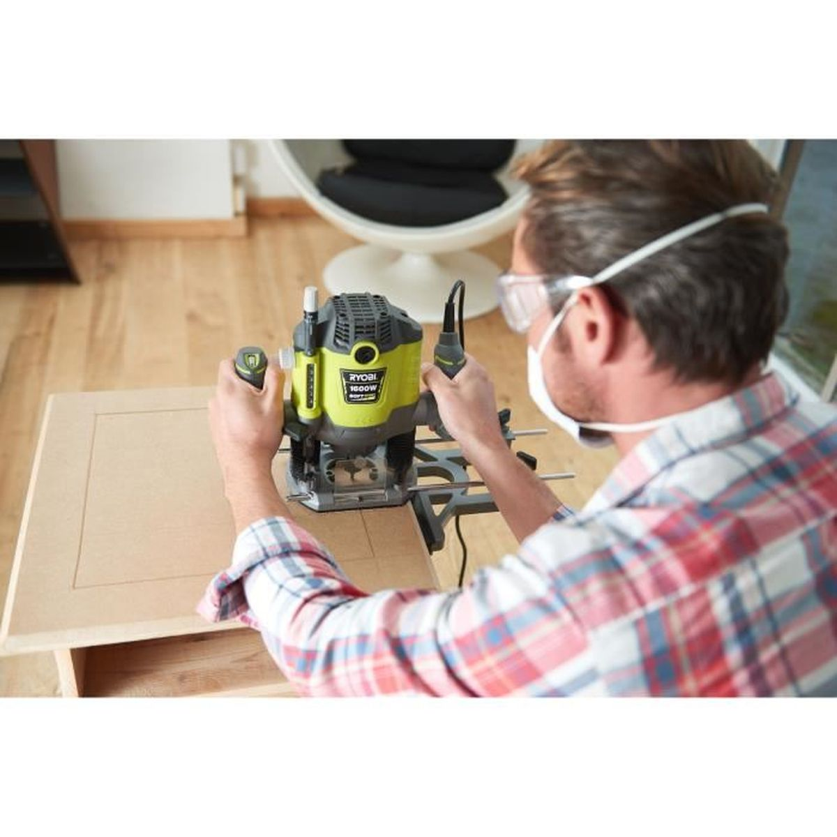 Mann mit Maske und Brille benutzt Ryobi-Fräser auf Holz. Von hinten gesehen. Holztisch, Boden und Stuhl.