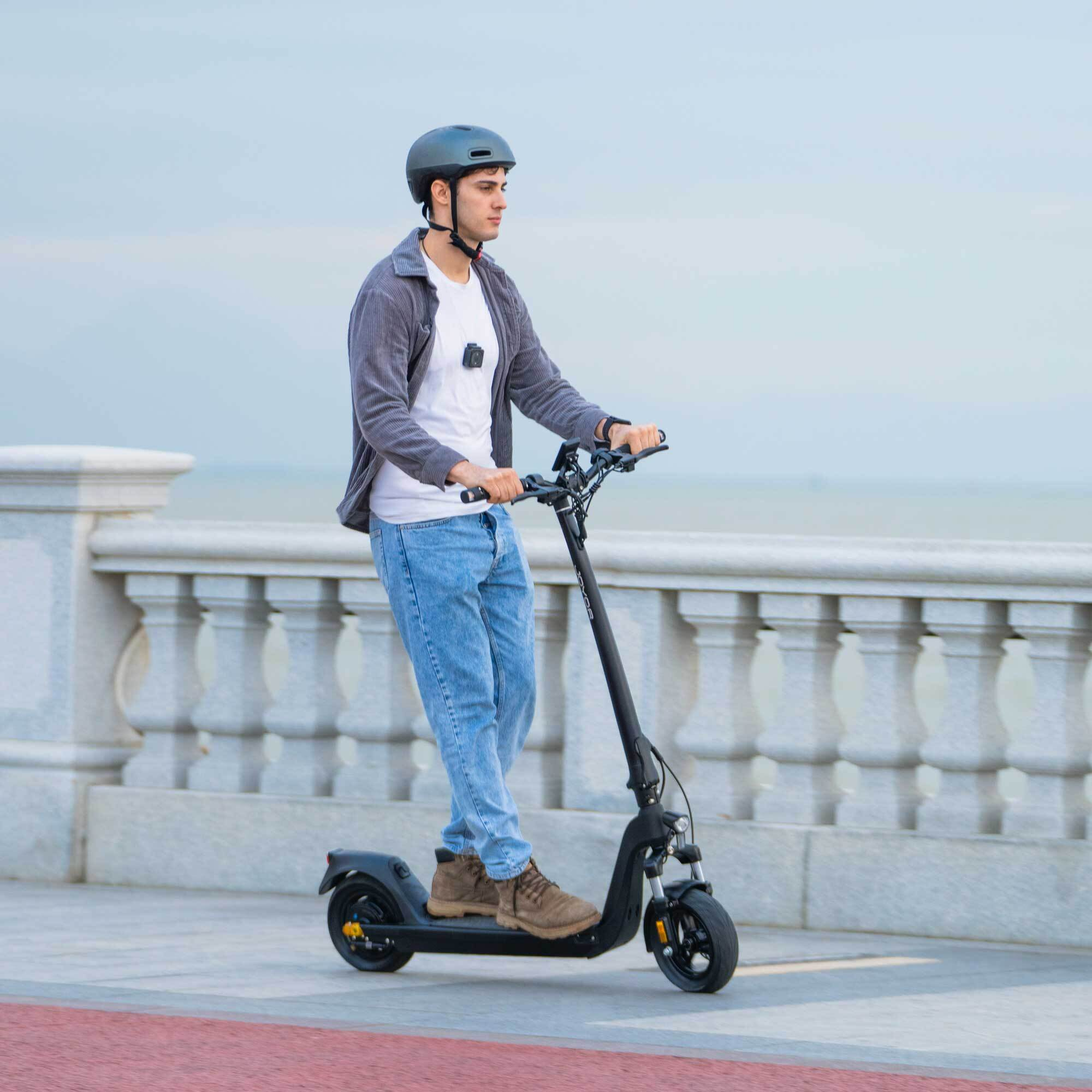 Un hombre en un patinete eléctrico negro, con casco, montado sobre una superficie pavimentada.