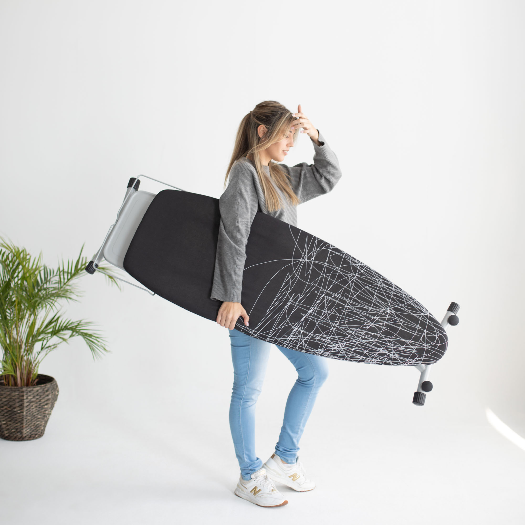 Mujer cargando una tabla de planchar, superficie blanca y planta atrás.