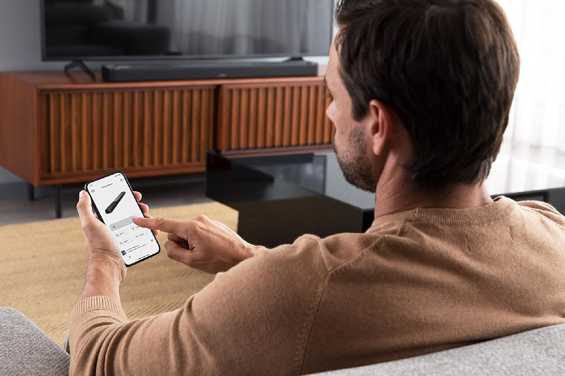 Ein Mann auf einem Sofa, der ein Telefon hält und mit dem Bildschirm interagiert. Eine Soundbar im Hintergrund.