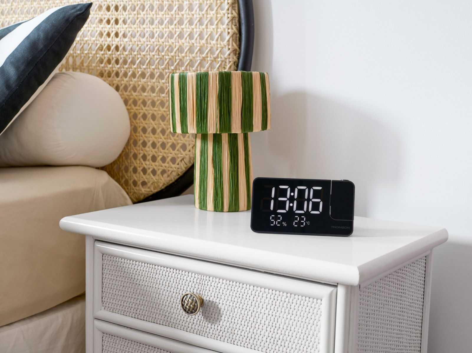Reloj despertador digital negro en una mesita de noche blanca, cerca de una lámpara y una cama.