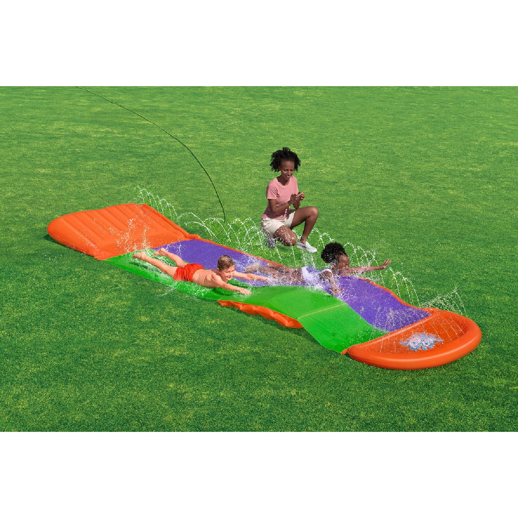 Drei Kinder auf einer Wasserrutsche, mit Wasserspritzern. Ein Kind sitzt auf dem Rasen und schaut zu.