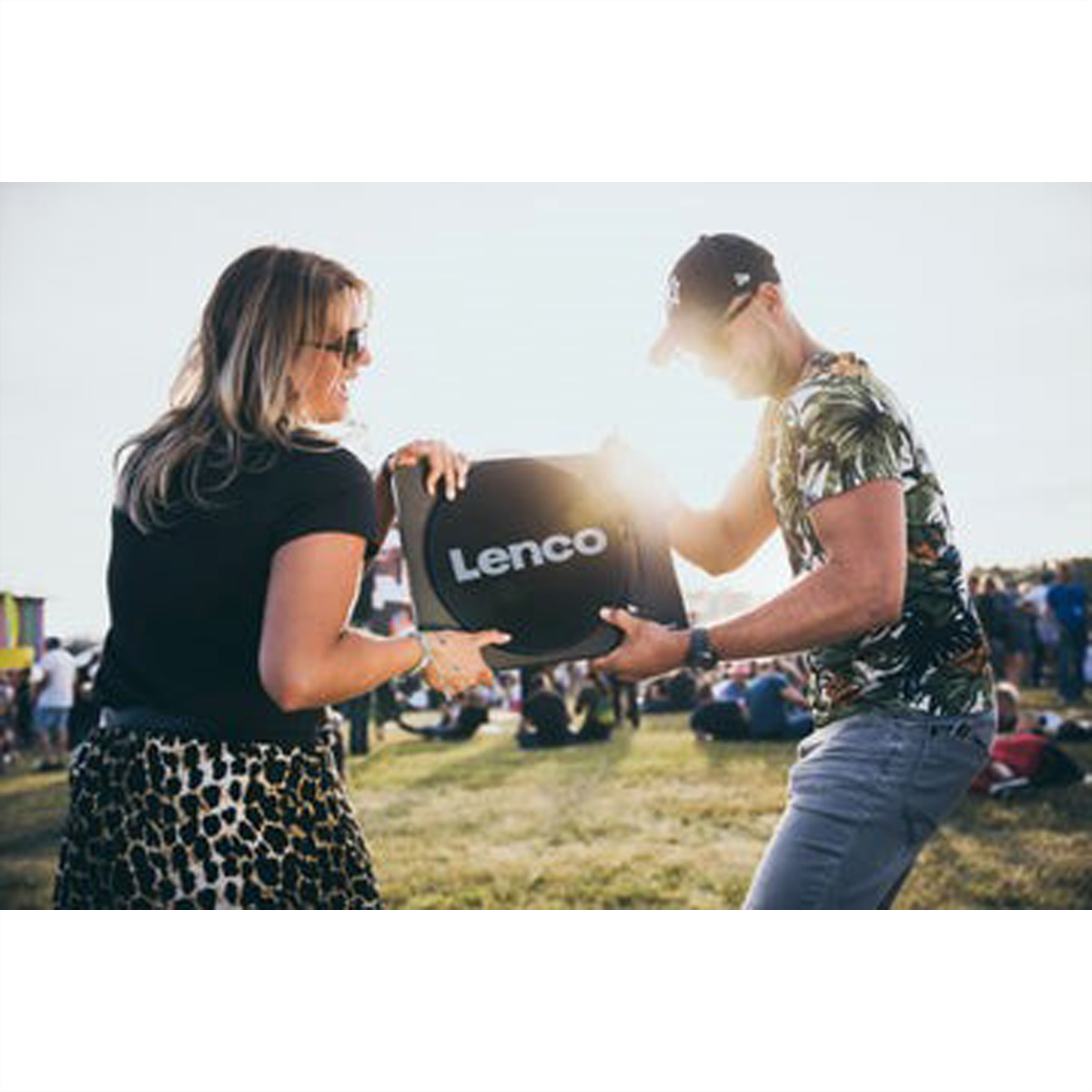 Dwie osoby trzymające czarny gramofon Lenco przed tłumem ludzi w świetle słonecznym.