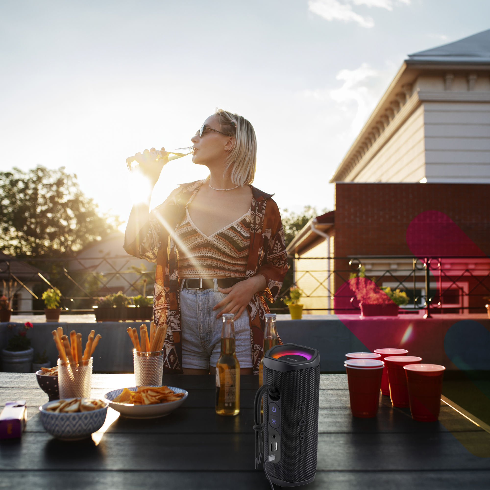 Osoba pijąca z butelki przy stoliku na zewnątrz z jedzeniem i głośnikiem. W tle zachód słońca.