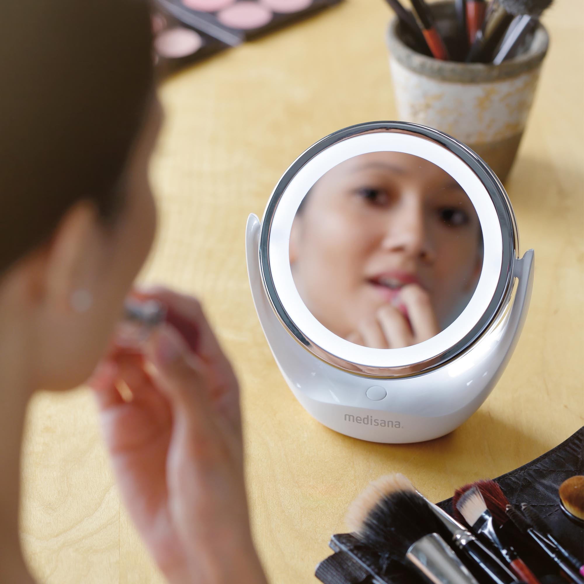 Donna che si guarda in uno specchio rotondo illuminato. Pennelli per trucco e cosmetici sul tavolo.