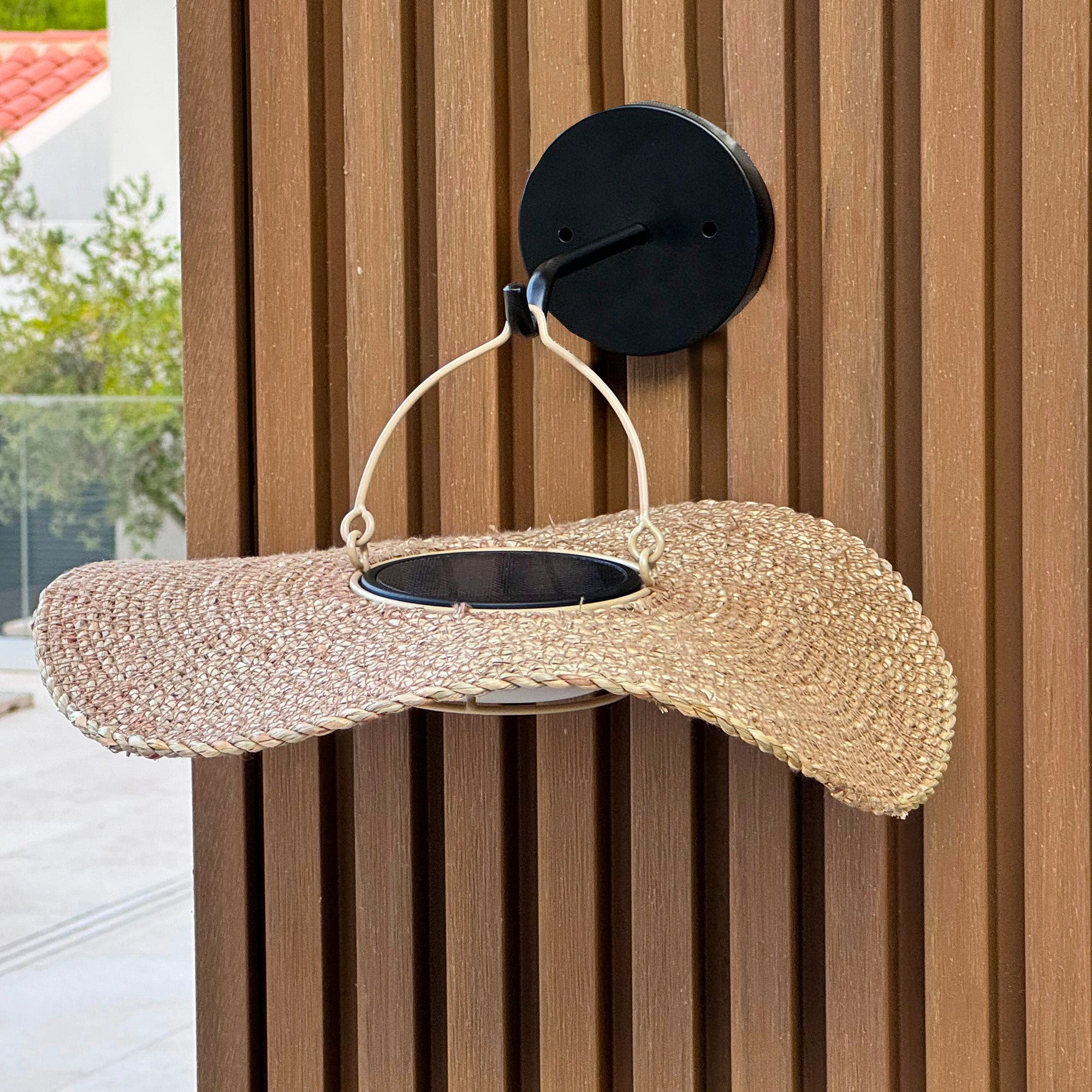 Ein gewebter Hut hängt an einem schwarzen Haken an einer Holzwand. Hintergrund zeigt einen Teil eines Hauses.