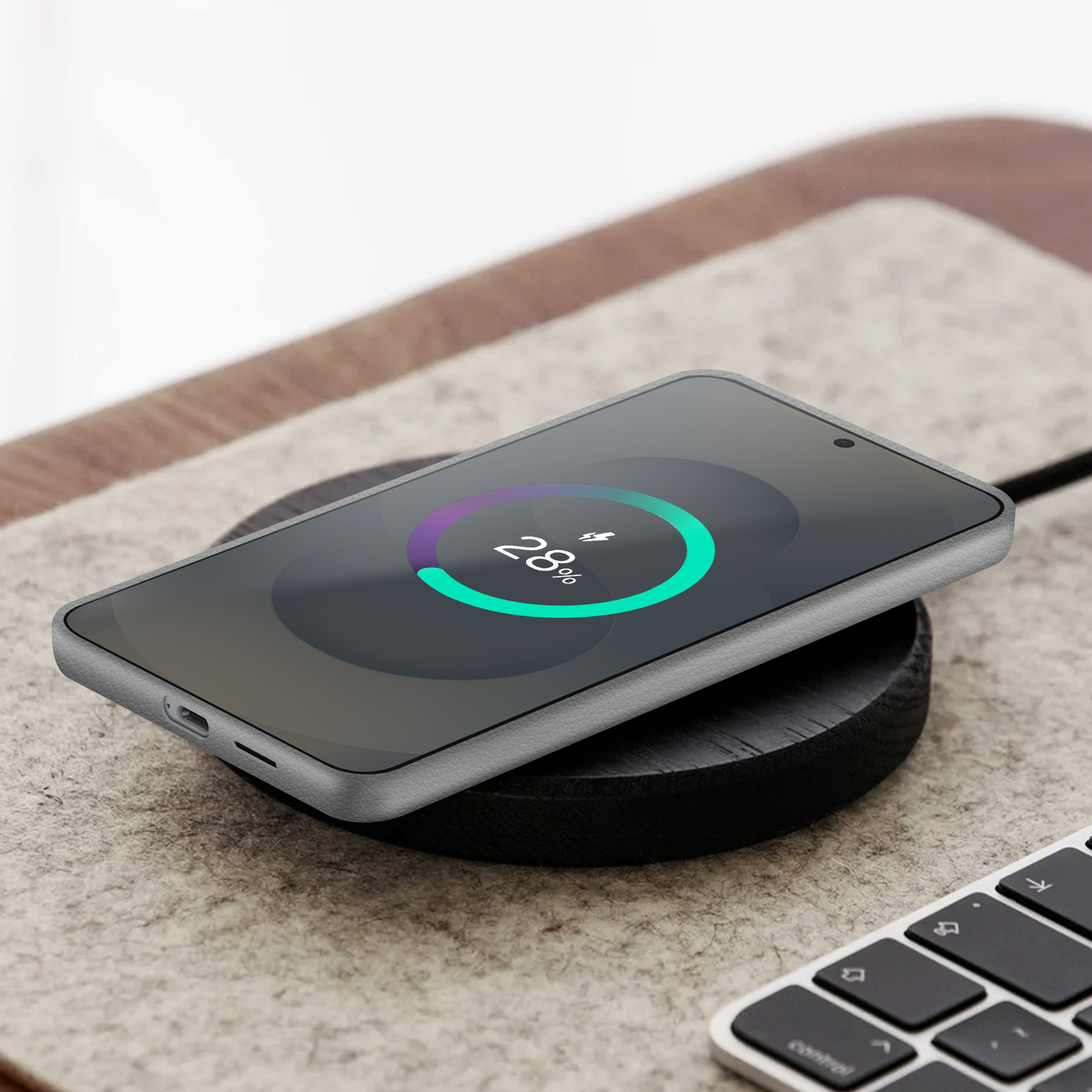 Smartfon na ładowarce bezprzewodowej pokazuje 28% naładowania. Ładowarka leży na filcowej podkładce, w rogu klawiatura.
