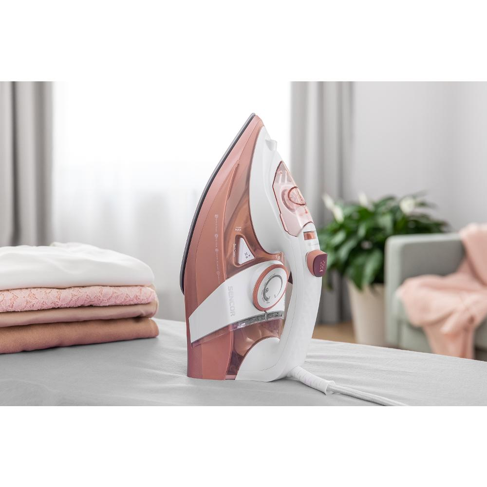 Un ferro da stiro, con un design bianco e rosa, su un'asse da stiro grigia con vestiti piegati.