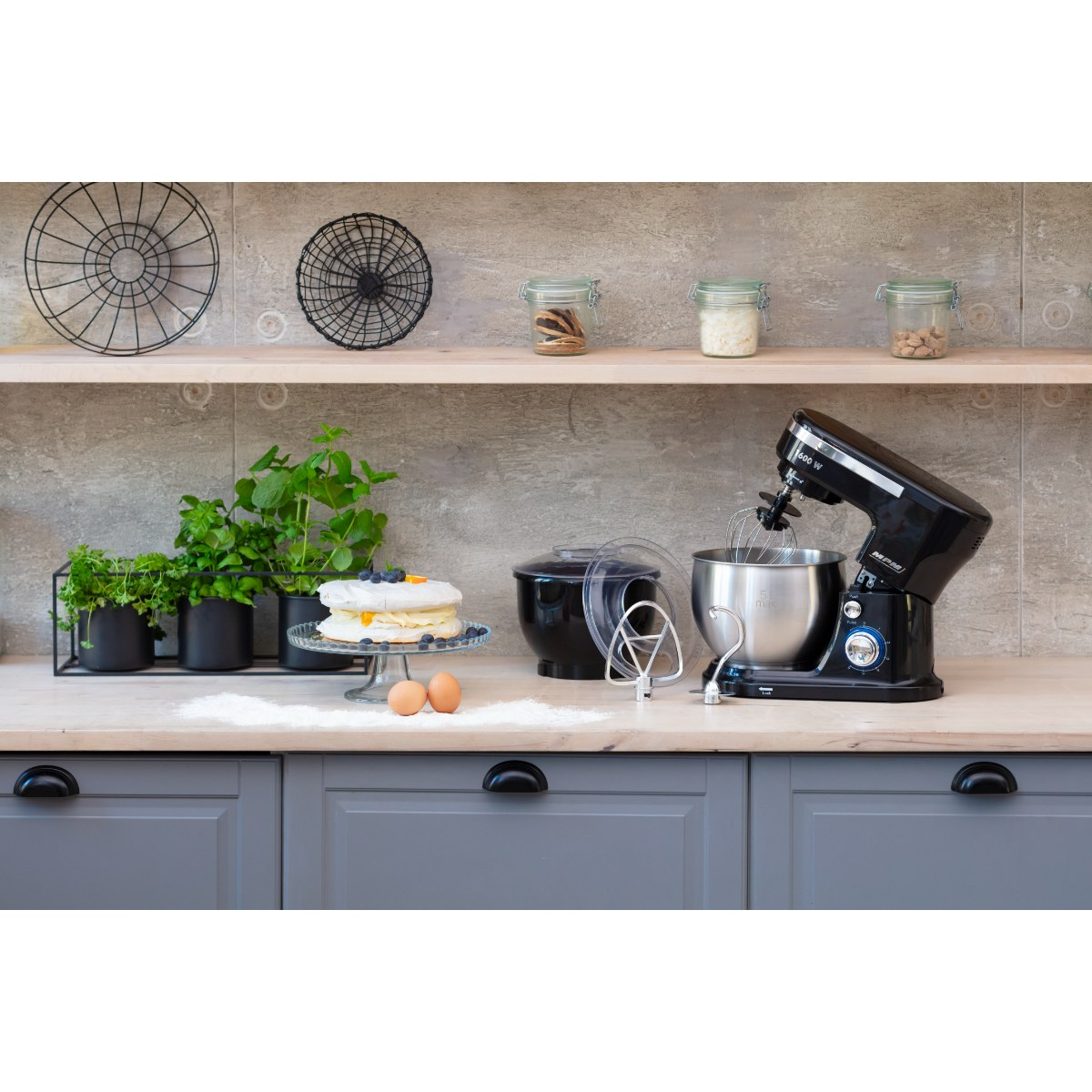 Scena na blacie kuchennym z czarnym mikserem, ciastem, ziołami i słoikami.