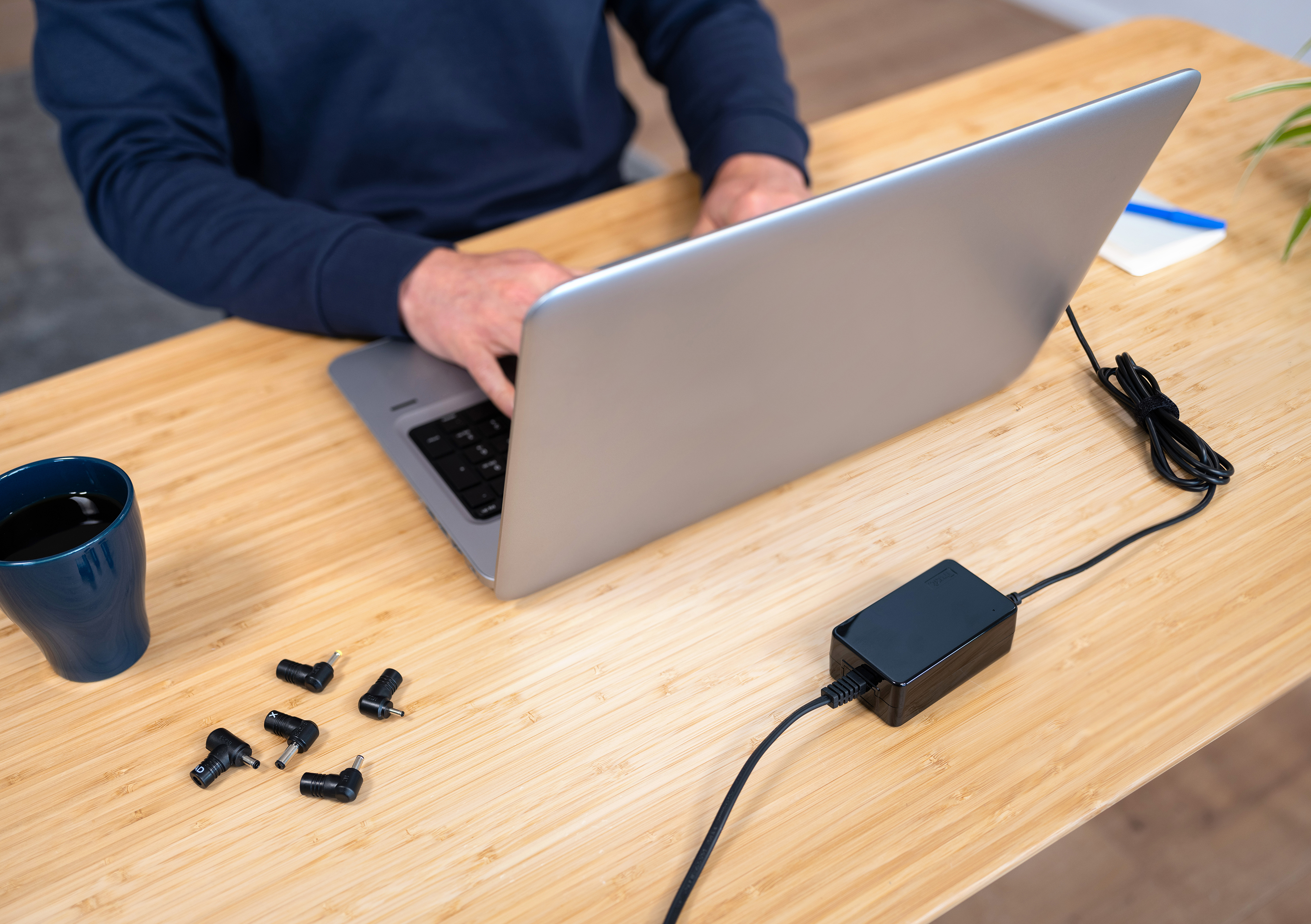 Person tippt auf Laptop, schwarzer Netzadapter und Adapterköpfe auf Schreibtisch.