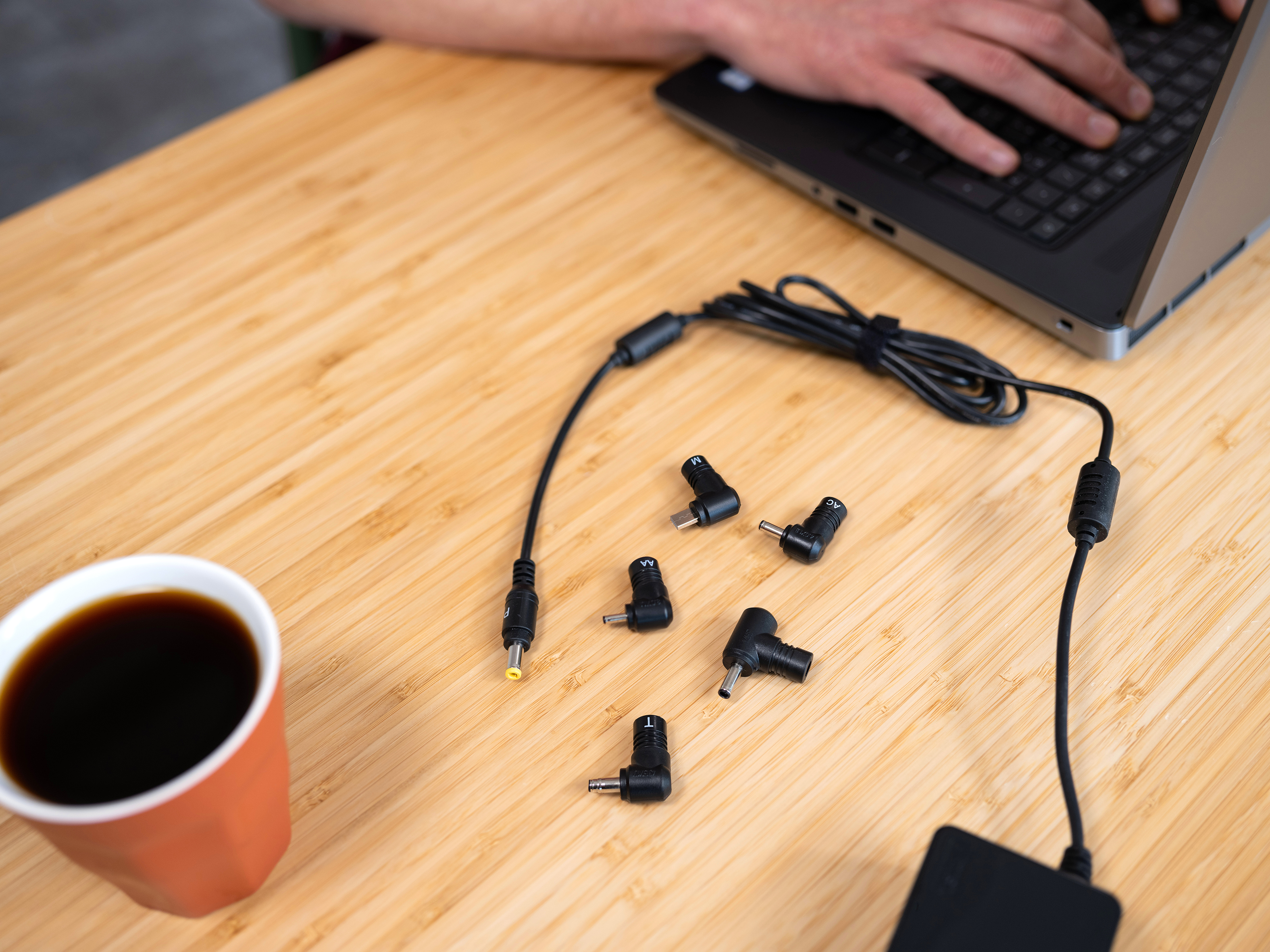 Laptop with power adapter and various tips on a wooden table. Coffee cup is on the left.