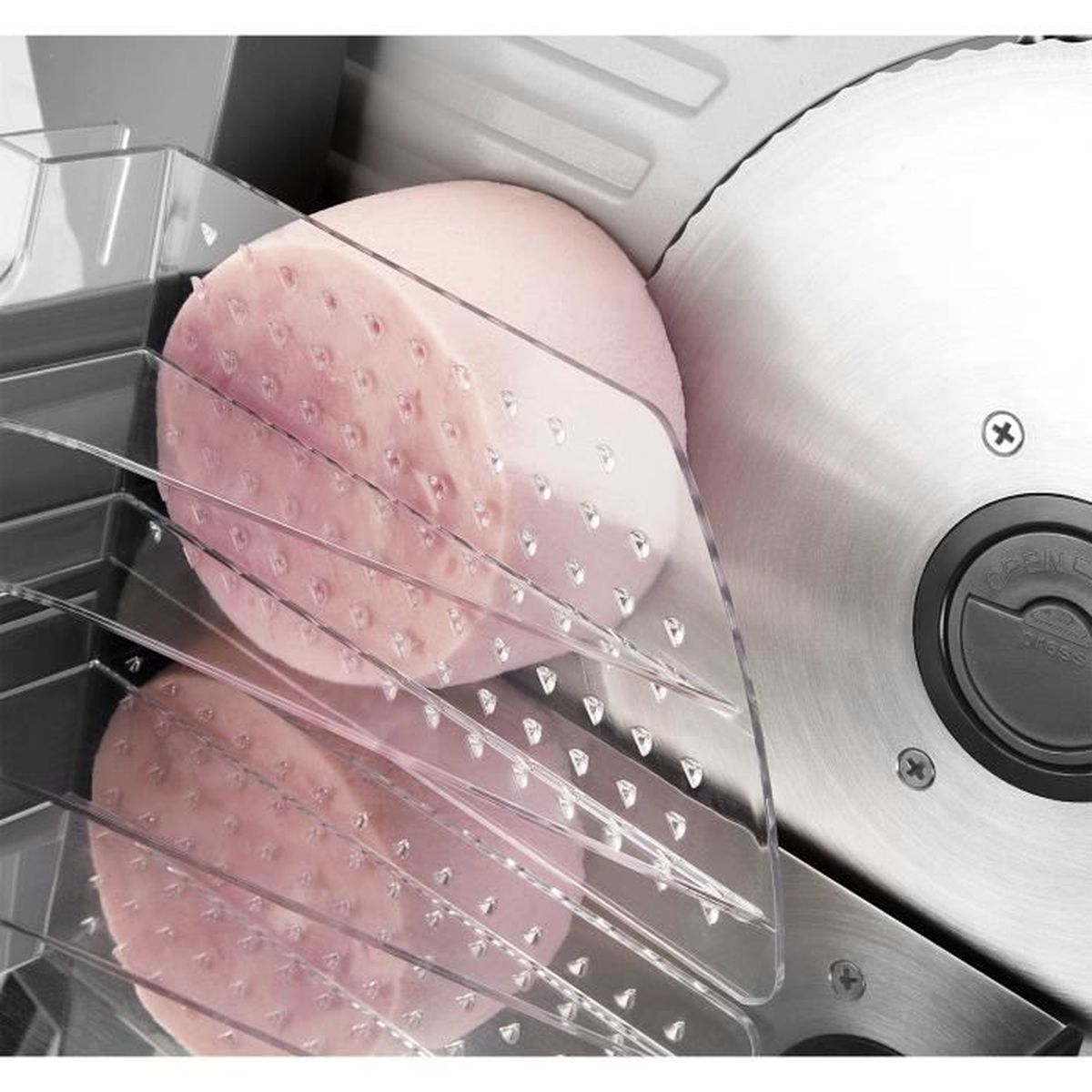 Zbliżenie krajalnicy do mięsa z kawałkiem różowej szynki w trakcie krojenia. Przezroczysta plastikowa osłona.