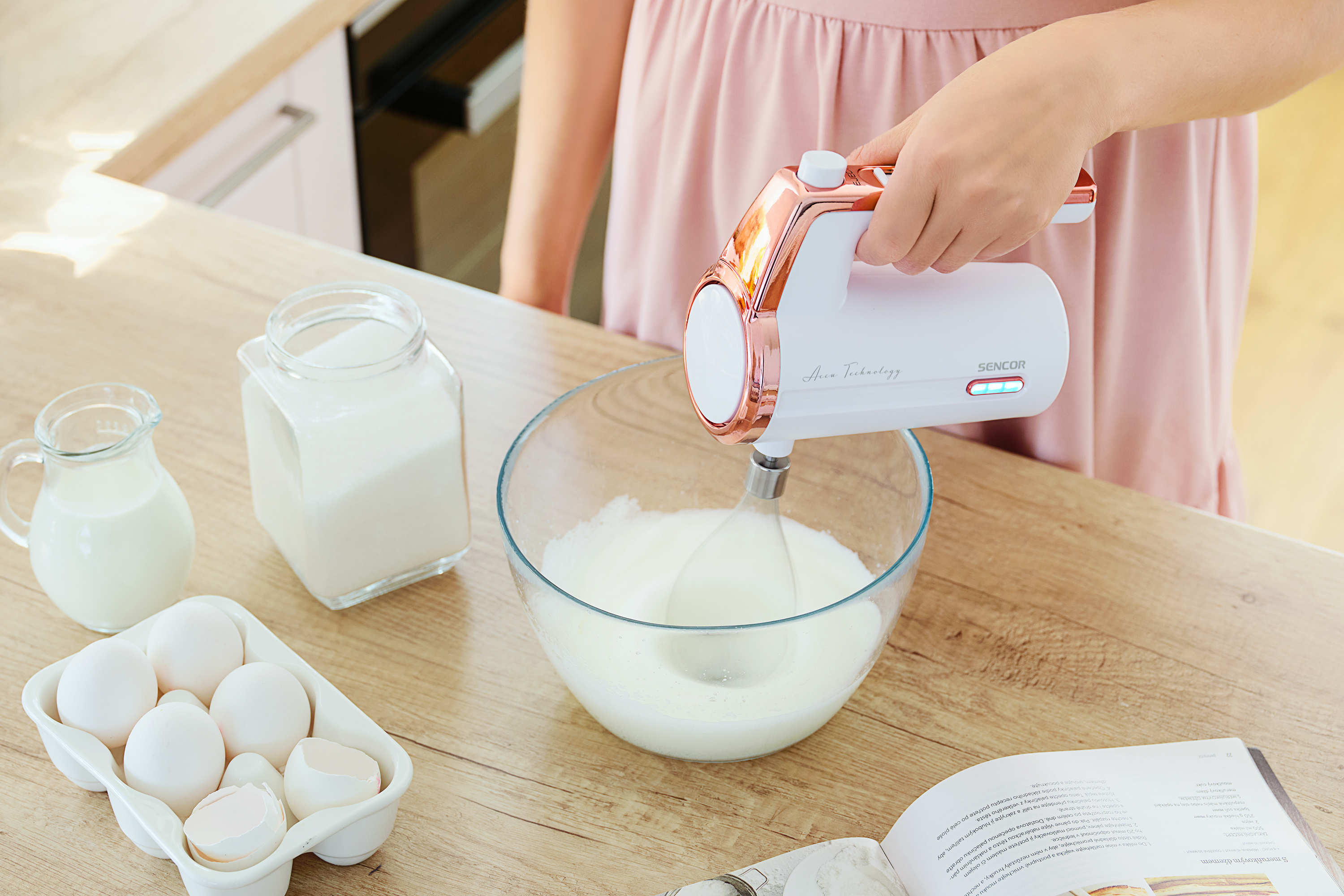 Una donna usa un frullatore a mano bianco e oro rosa per sbattere gli ingredienti in una ciotola di vetro.