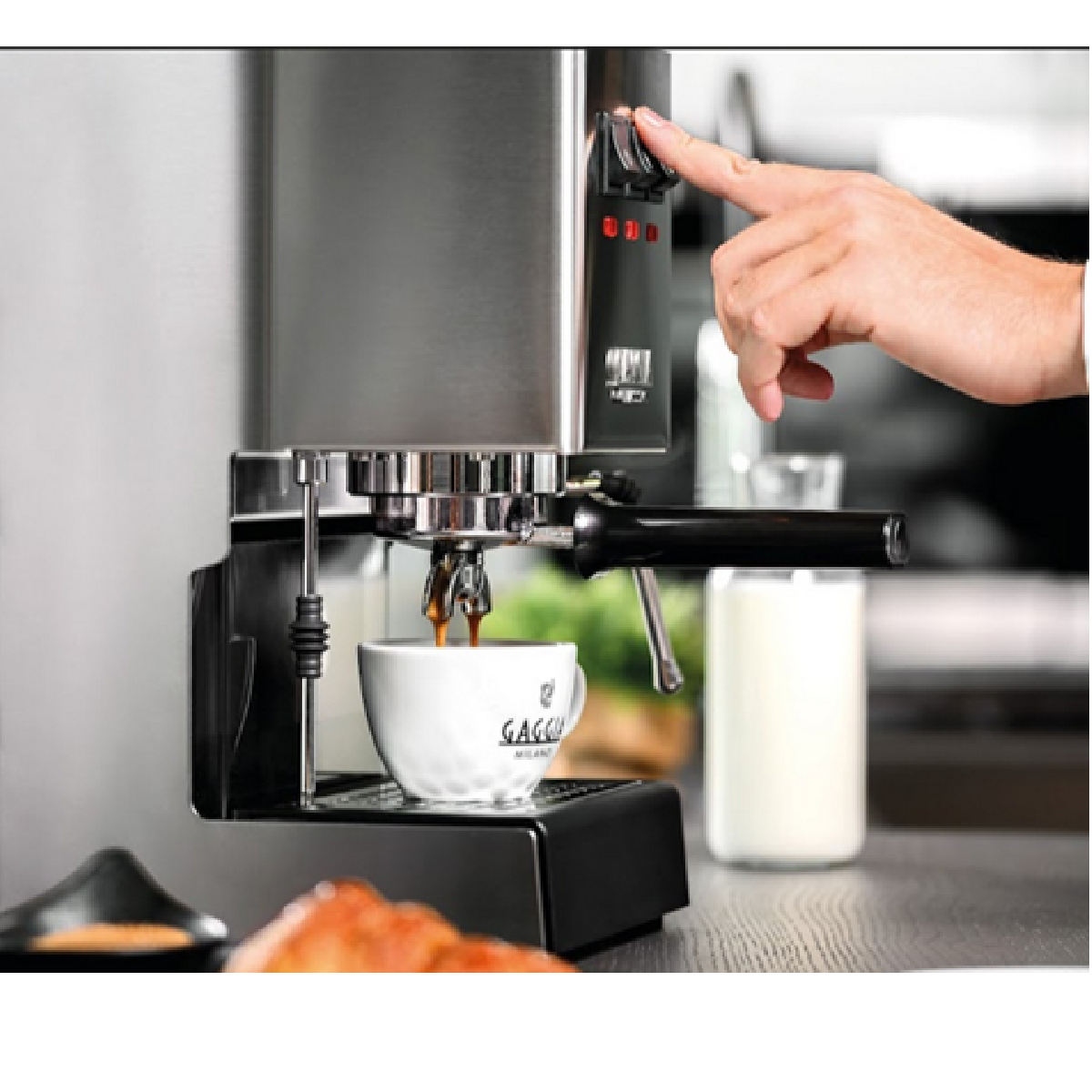 Espressomachine met een kopje, koffie die schenkt. Hand drukt op een knop. Melkglas en croissant zichtbaar.