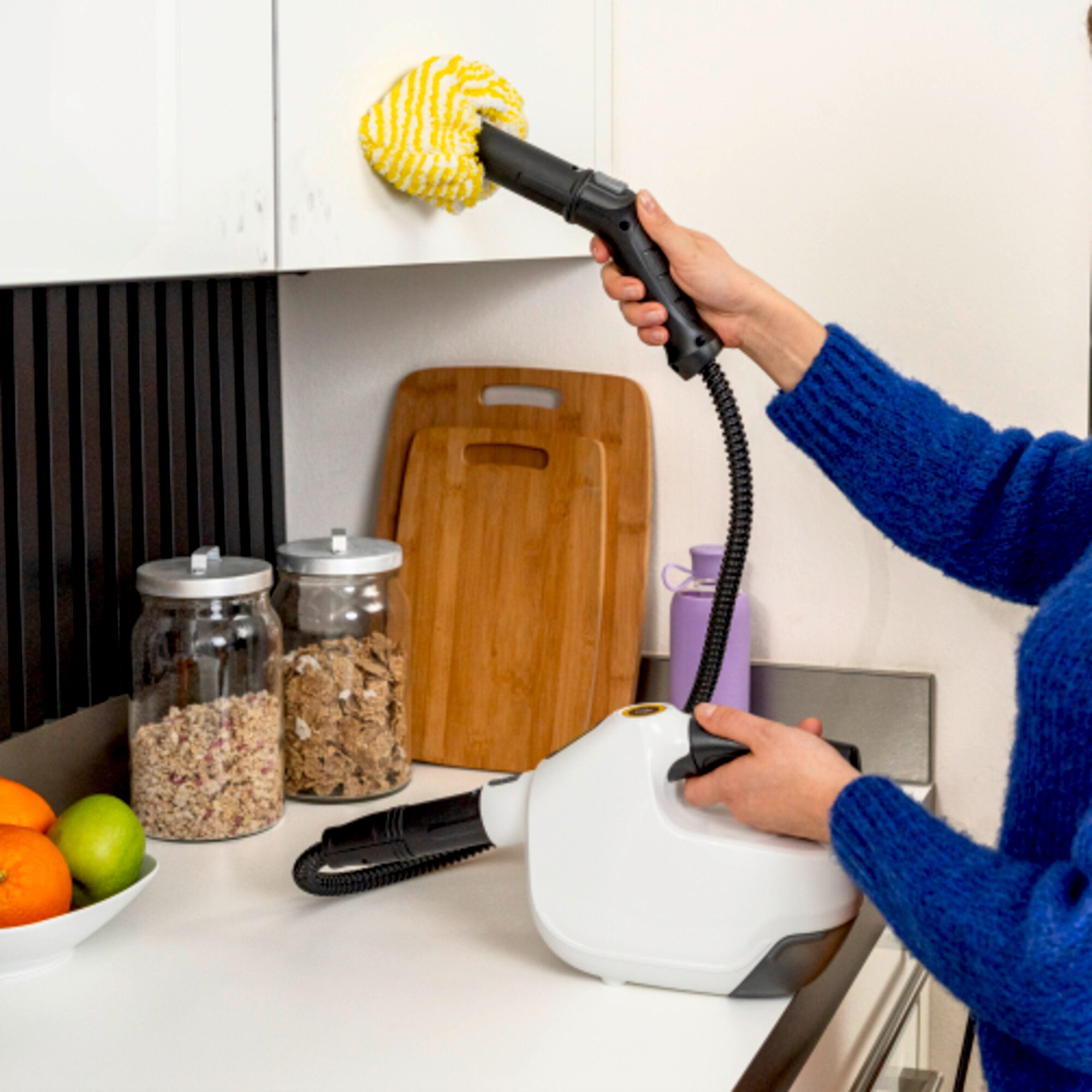Osoba czyszcząca białą szafkę parownicą. Tło kuchni z deskami, słoikami i owocami.