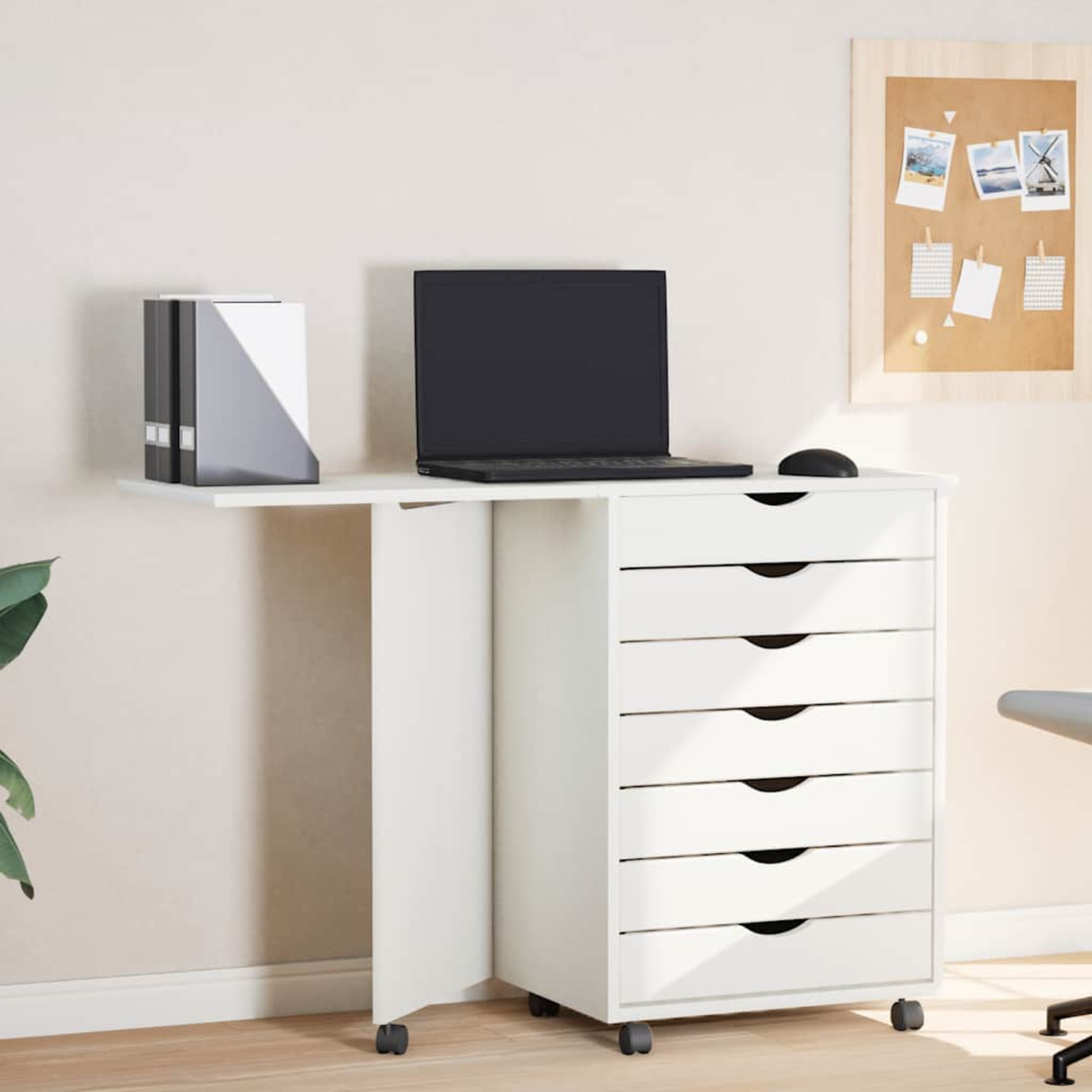 Weißer Schreibtisch mit Laptop, Schubladen und einer Pinnwand mit Fotos, auf Rädern, neben einer Wand.