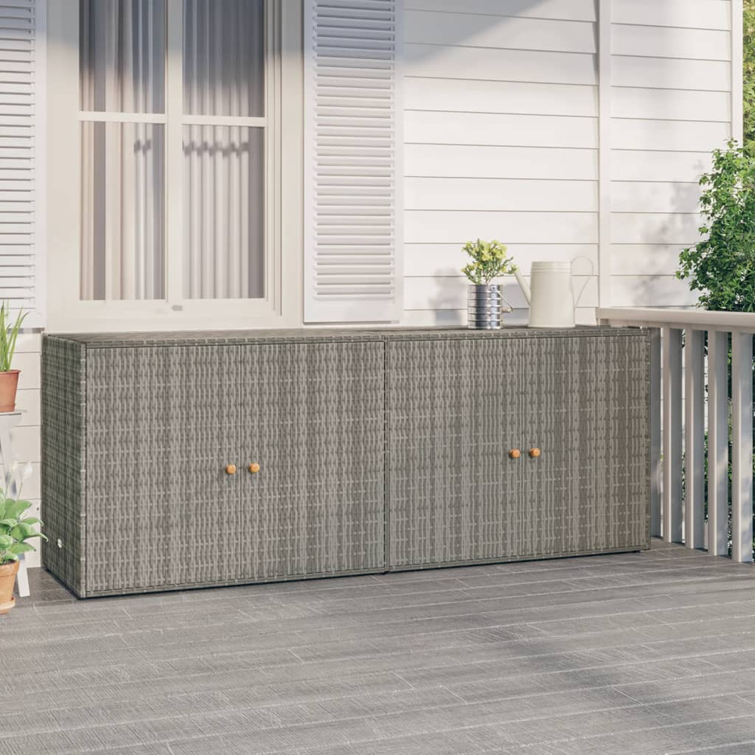 Grauer Rattan-Schrank mit Holzgriffen, auf einer Veranda. Ein weißes Fenster und Pflanzen sind sichtbar.