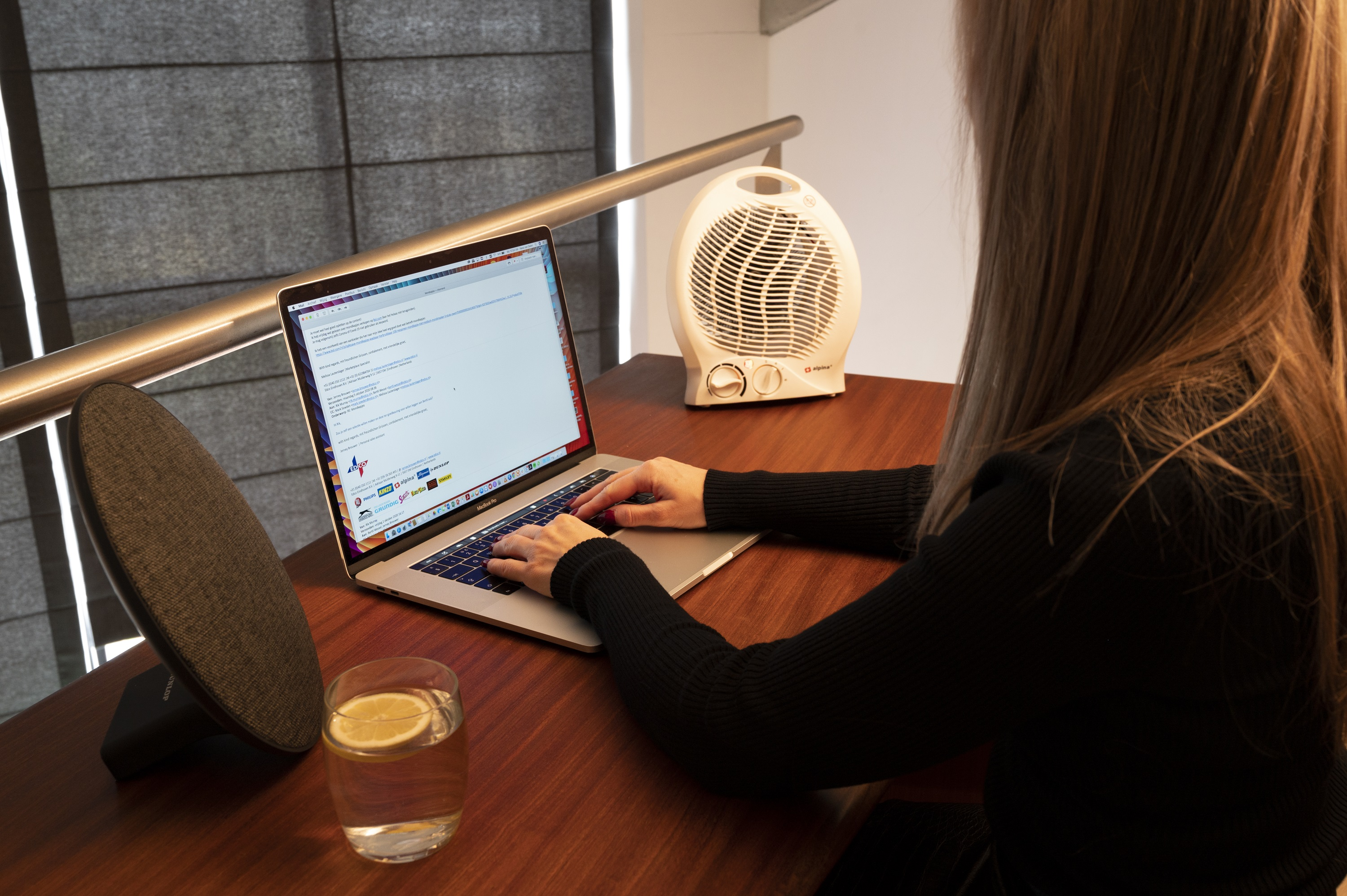 Een vrouw werkt op een laptop. Er staat een luidspreker en een glas water op de tafel.