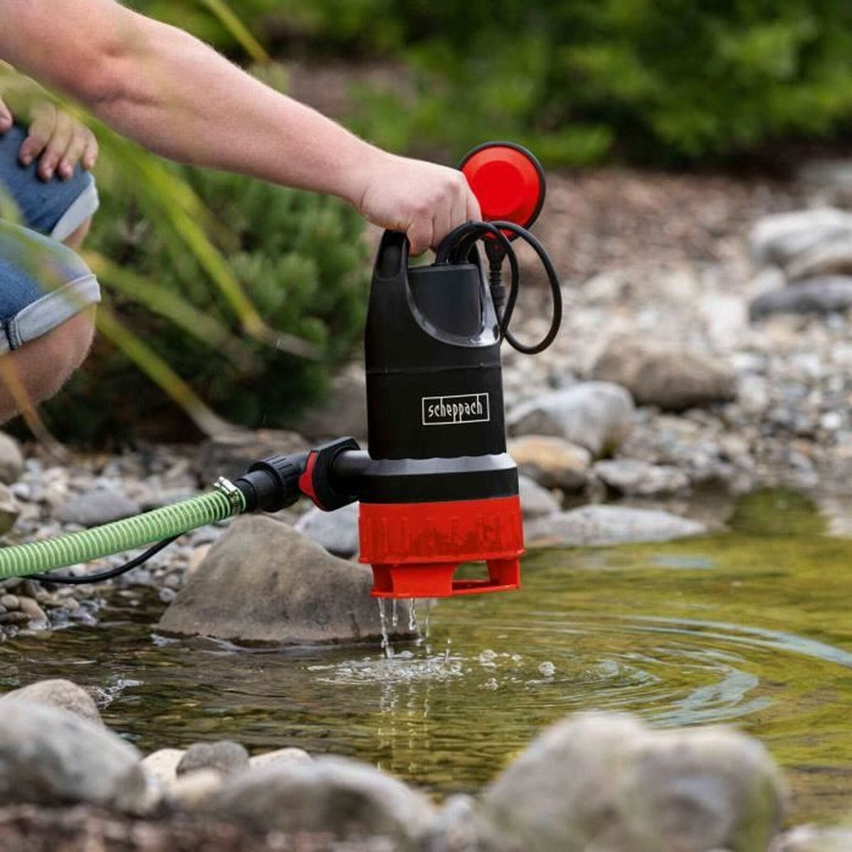 Eine Person hält eine schwarz-rote Wasserpumpe vor einem Teich. Grüner Schlauch angeschlossen.