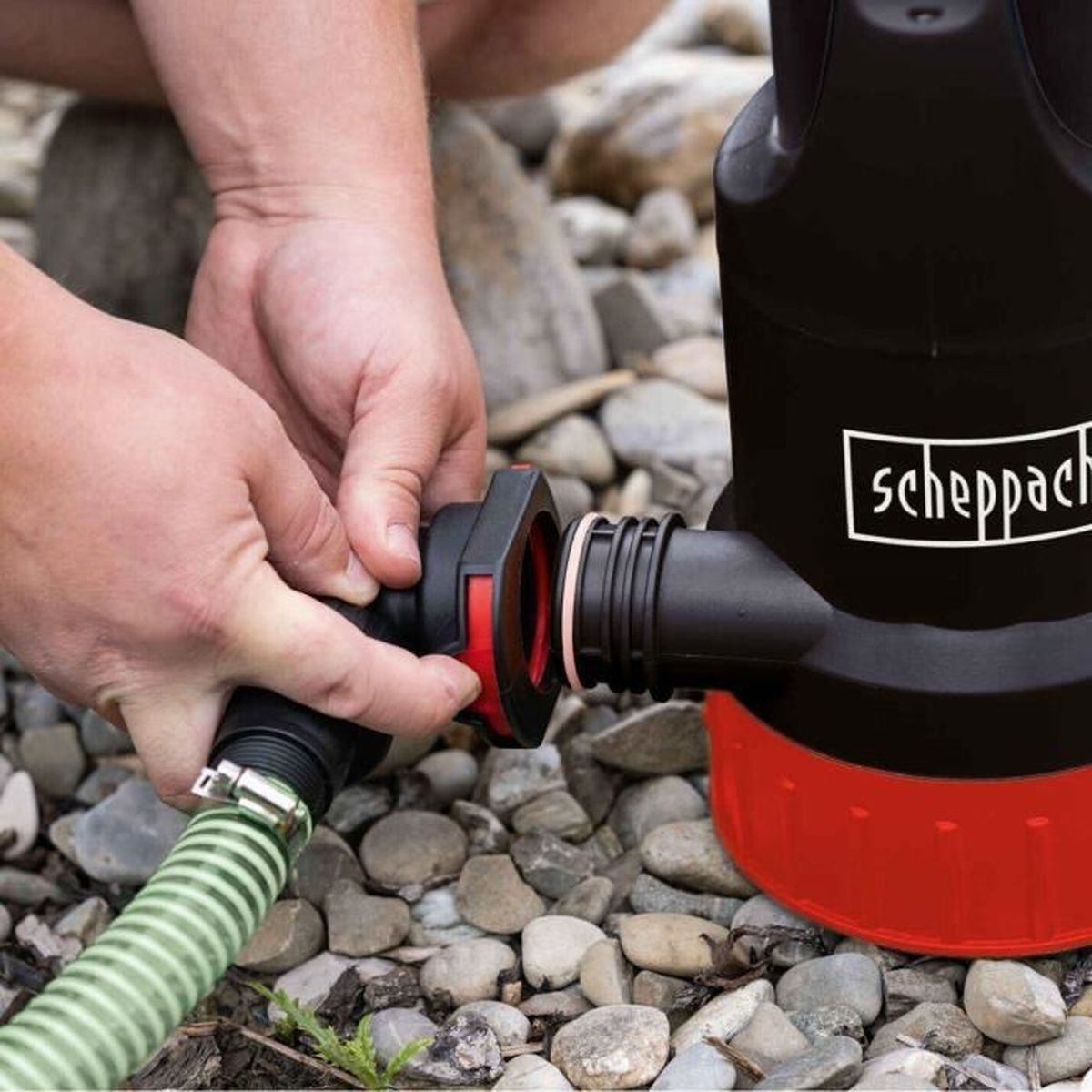 Ein Mann verbindet einen grünen Schlauch mit einer schwarz-roten Wasserpumpe auf steinigem Untergrund.