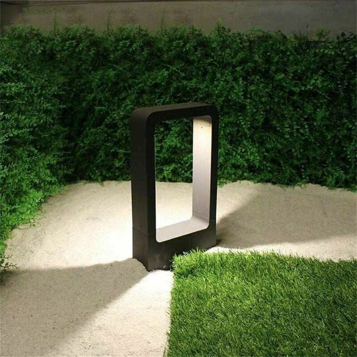 Schwarze, rechteckige Außenlampe auf einem weißen Steinweg, begrenzt von Gras und grünen Büschen.