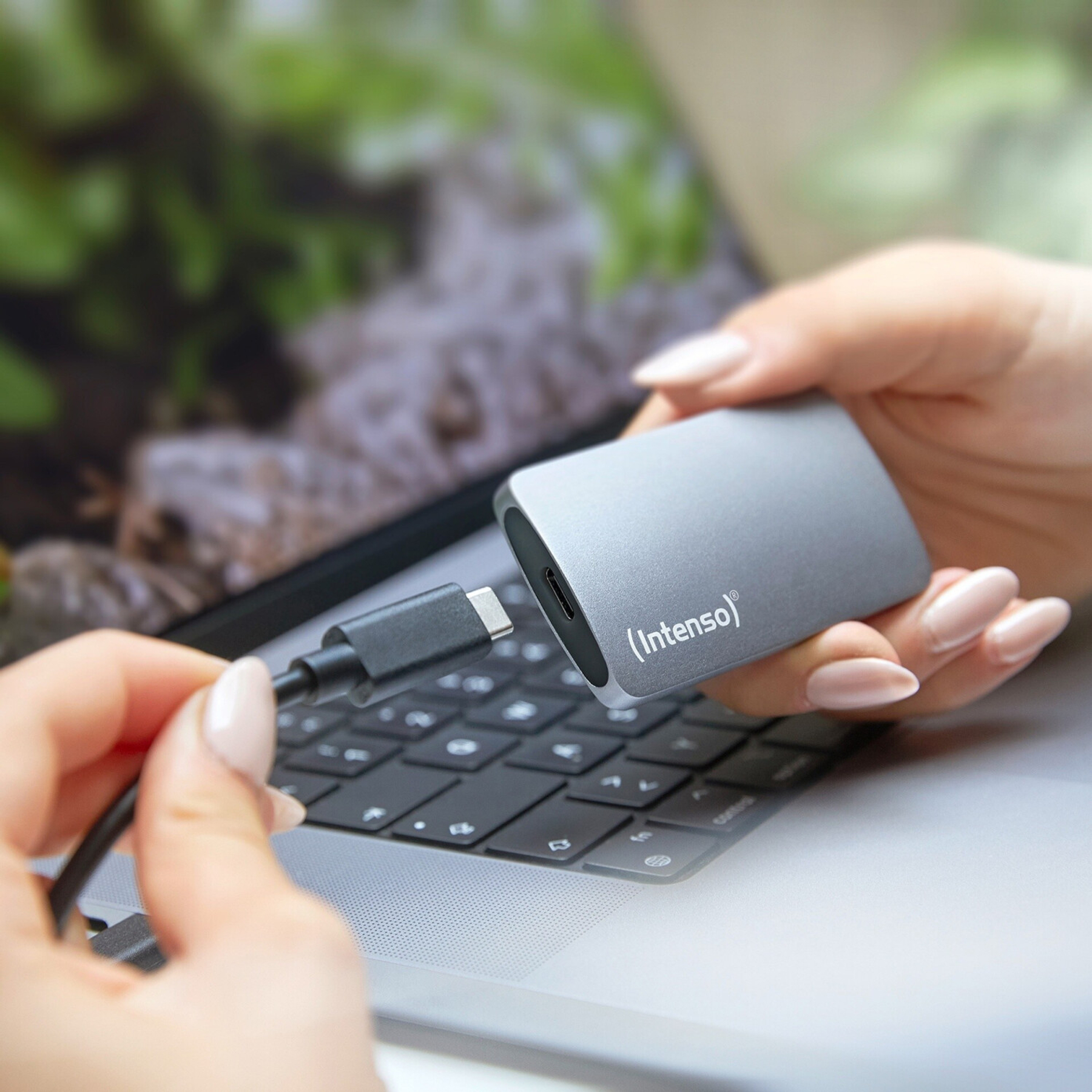 Osoba trzymająca szary zewnętrzny dysk twardy w pobliżu laptopa z kablem ładującym.