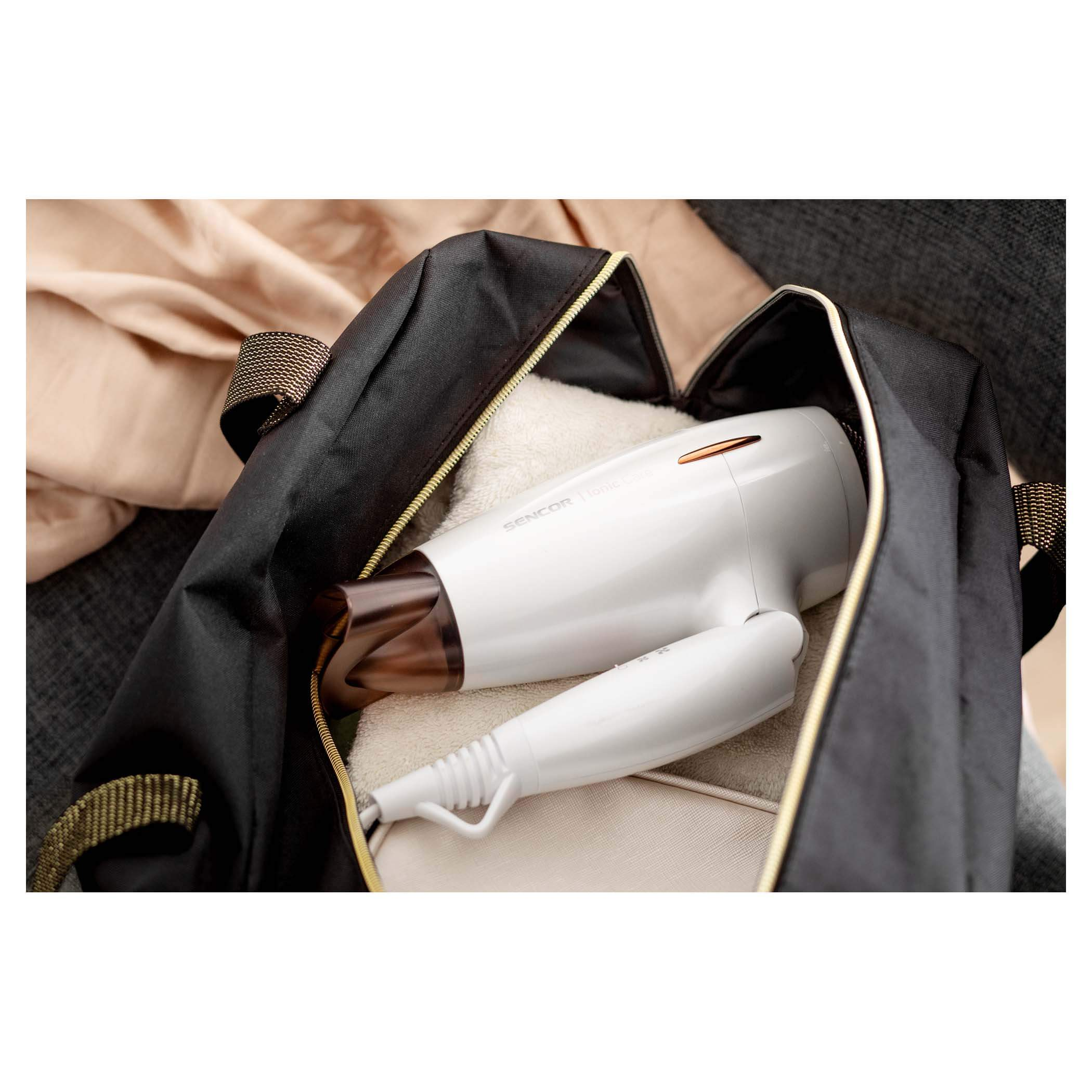 Czarna torba sportowa ze złotymi akcentami. Biała suszarka do włosów w środku. Beżowa powierzchnia w tle.