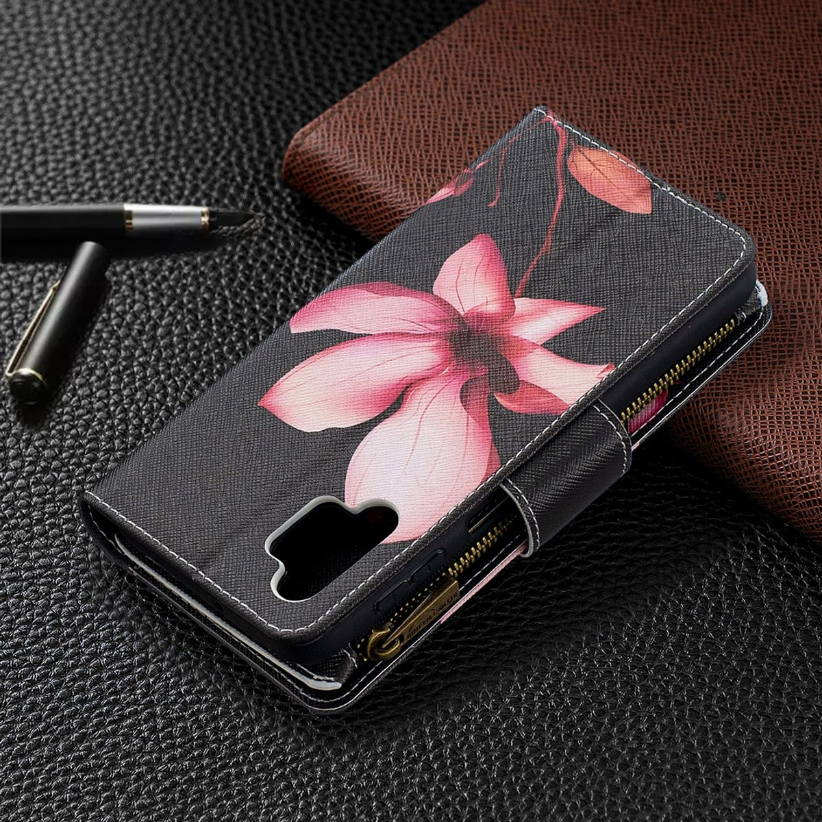 Schwarze Handyhülle mit rosa Blumendesign, geöffnet auf strukturierter Oberfläche. Goldener Reißverschluss und Stift.