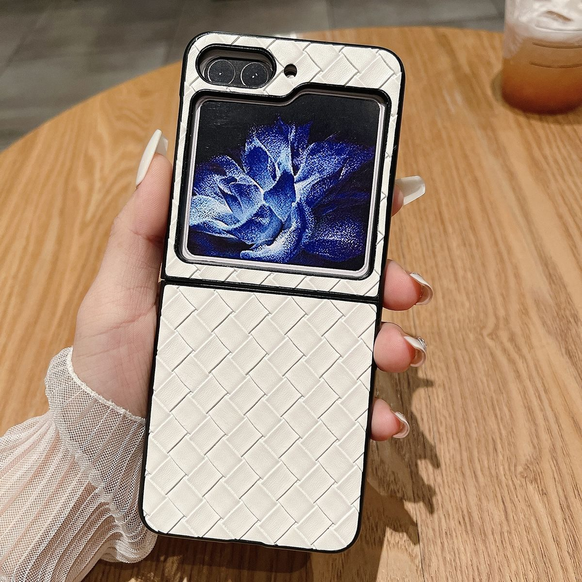 Eine weiße, gewebte Handyhülle, gehalten von einer Hand. Durch die Öffnung ist ein blaues Blumenbild zu sehen.