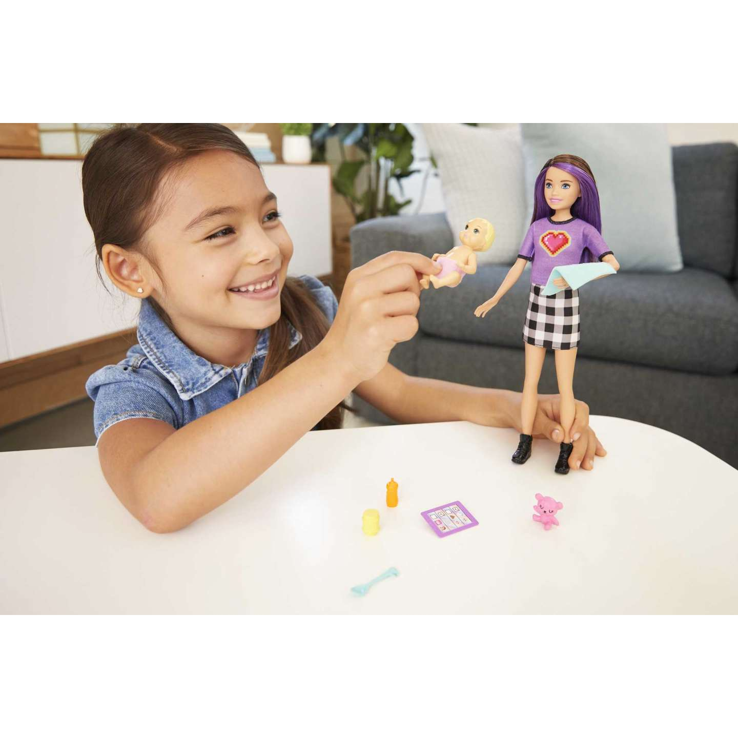 Una niña sonriendo, sosteniendo una muñeca bebé. Una muñeca Barbie y accesorios están en la mesa.