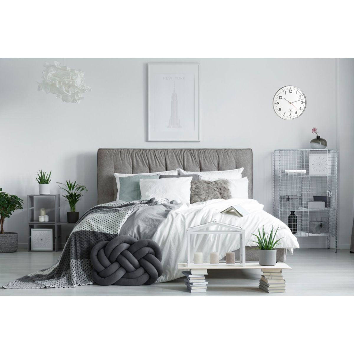 Sypialnia z łóżkiem, poduszkami i dekoracjami. Widoczny jest zegar i oprawiony wydruk.