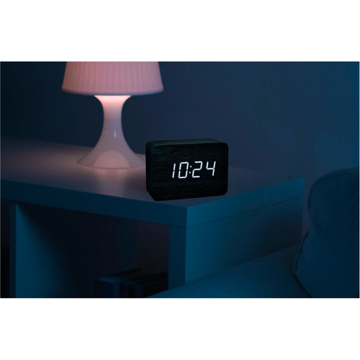 Schwarze Holzuhr auf einem weißen Tisch. Die Uhr zeigt 10:24 an. Eine Lampe auf der linken Seite.