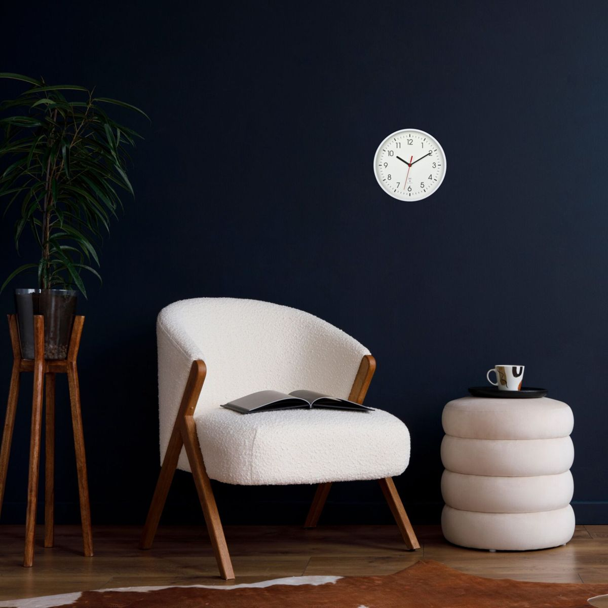 Ein weißer Stuhl, ein Tisch mit Tasse und eine Uhr an einer dunkelblauen Wand. Eine Pflanze links.