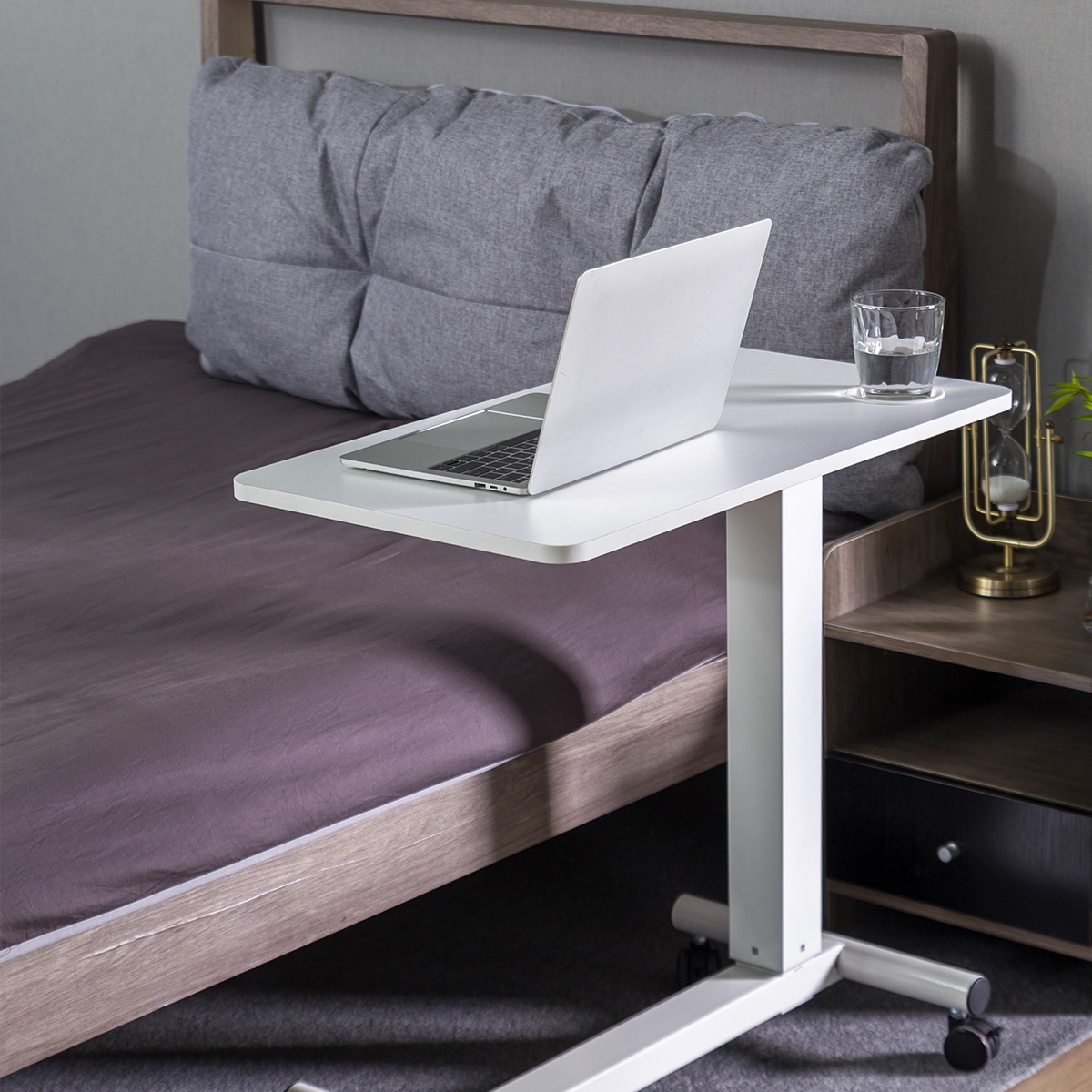 Ein Laptop steht auf einem weißen Tisch über einem Bett. Ein Glas Wasser steht auf dem Tisch. Eine Lampe und ein Nachttisch sind in der Nähe.