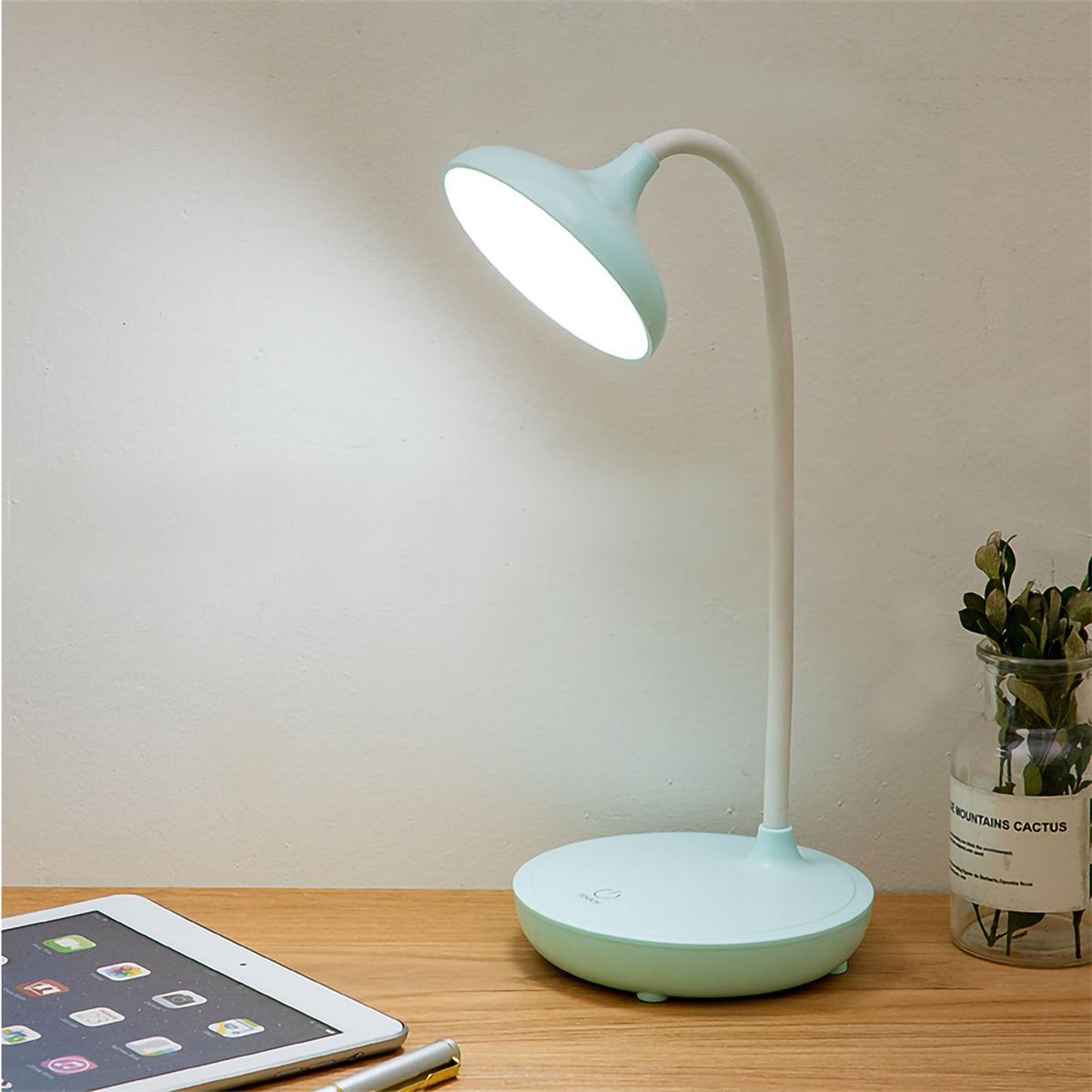 Eine mintgrüne Schreibtischlampe mit gebogenem Hals, beleuchtet, auf einem Holzschreibtisch.