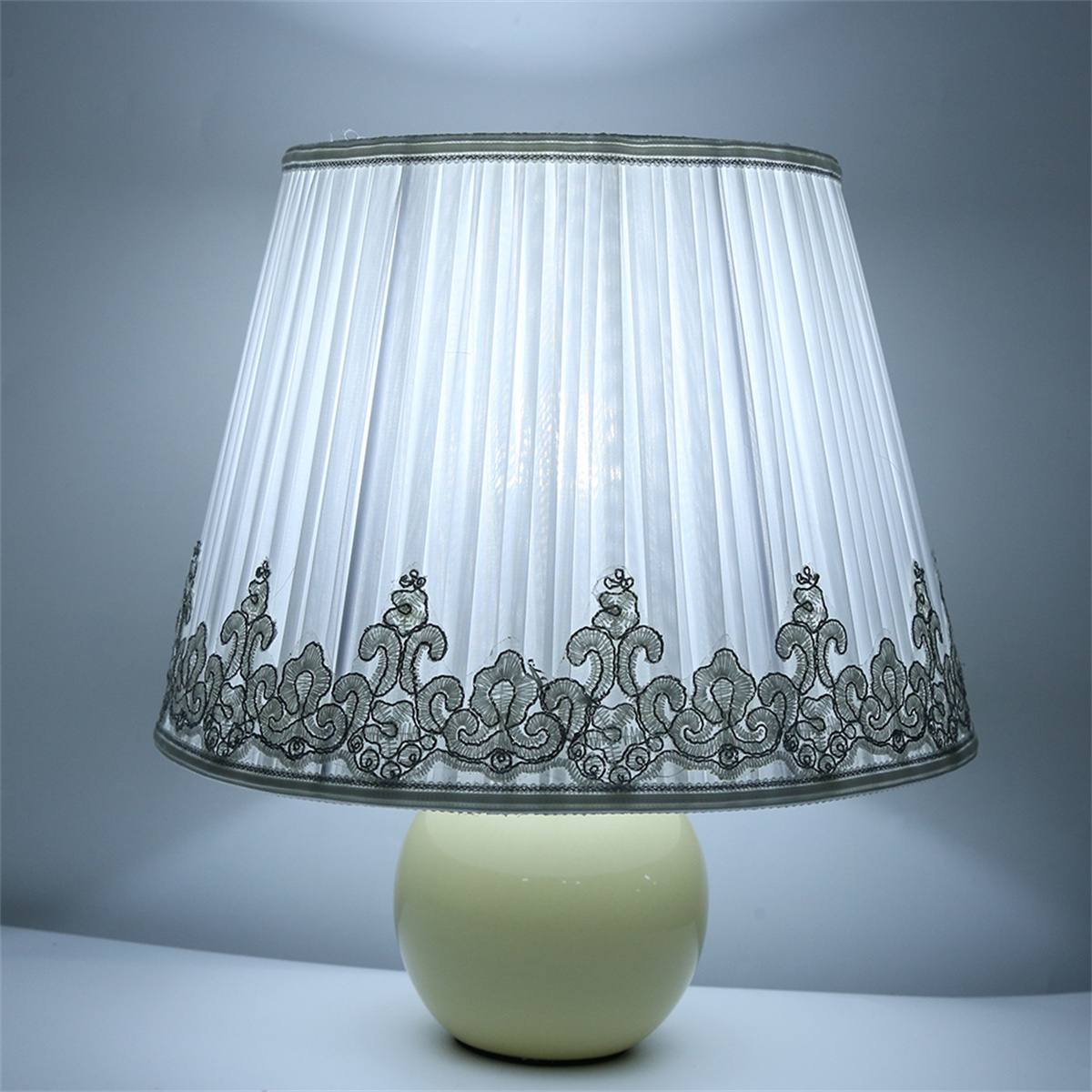 Eine cremefarbene Lampe mit weißem plissiertem Schirm und grauem Spitzensaum auf weißer Oberfläche.