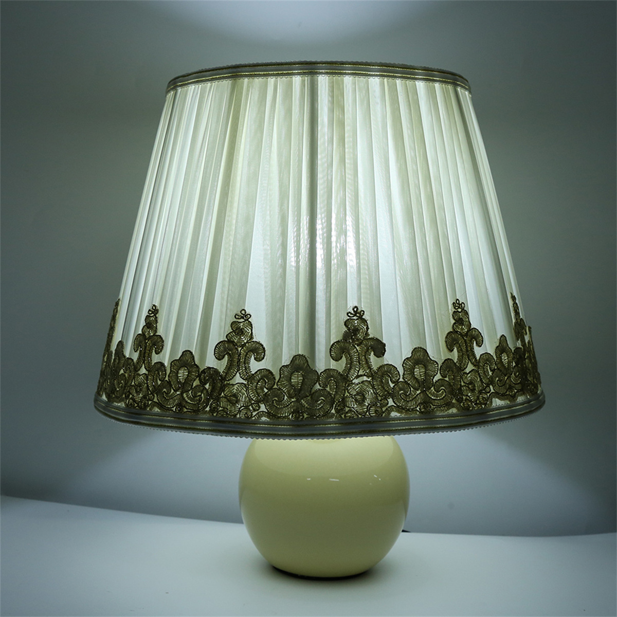 Eine cremefarbene Lampe mit einem plissierten Lampenschirm, der mit brauner Spitze verziert ist, auf einer weißen Oberfläche.