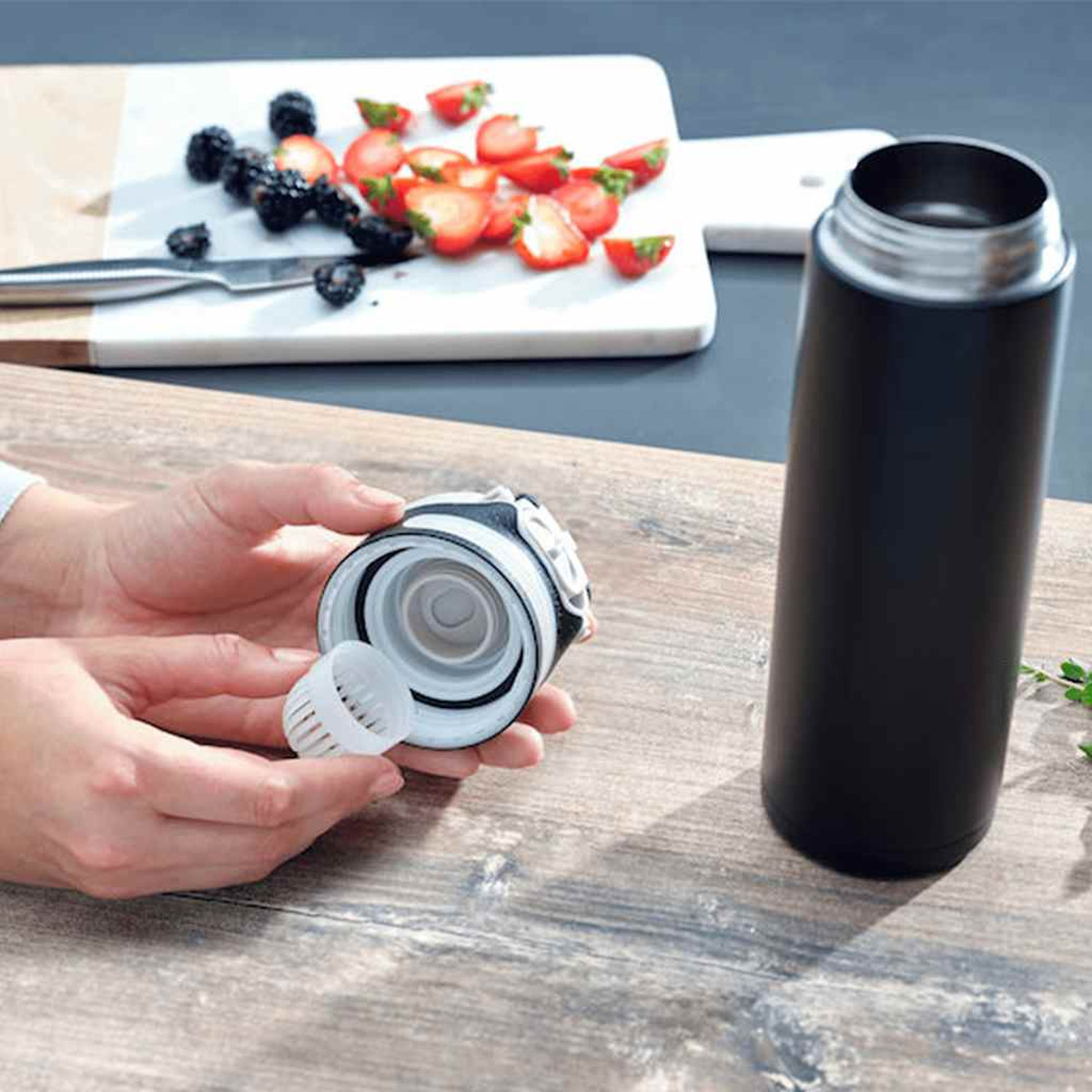 Eine Person hält eine Wasserflasche auseinander. Auf einem Schneidebrett liegen Erdbeeren und Brombeeren.