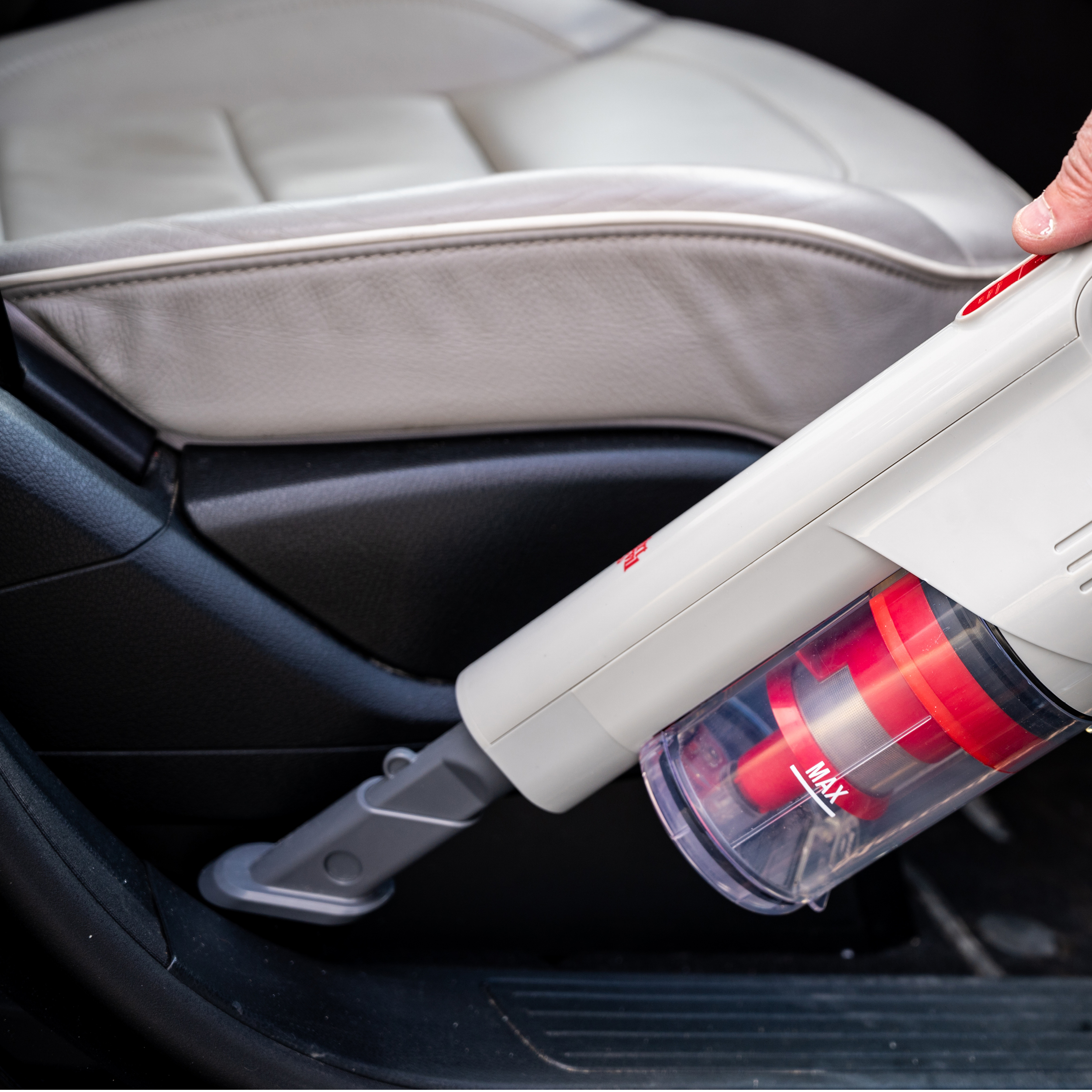 A hand using a handheld vacuum cleaner on a car seat. The vacuum cleaner is white.