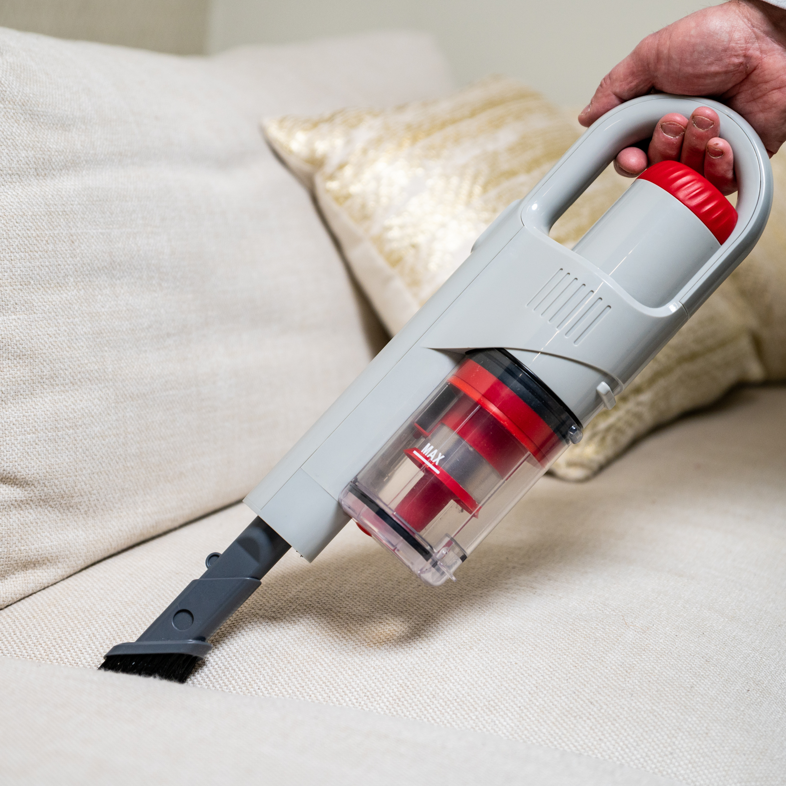 Hand holding a gray and red handheld vacuum cleaner, cleaning a white couch.