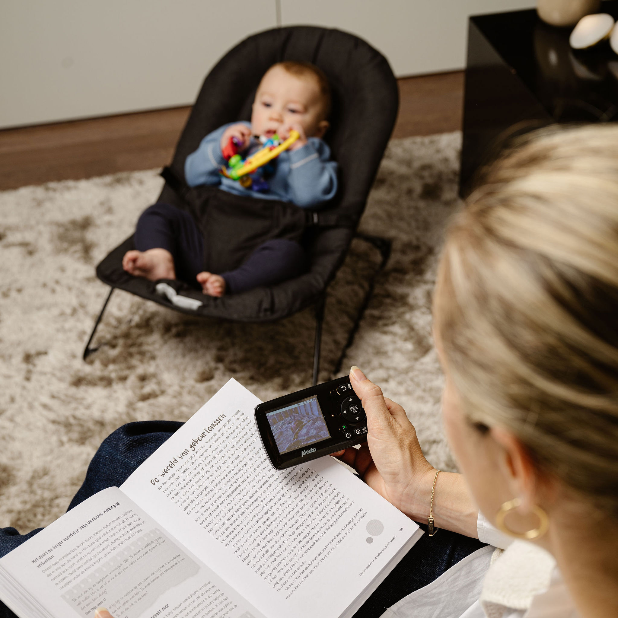 Matka czyta książkę trzymając nianię elektroniczną. Dziecko siedzi w leżaczku, bawiąc się zabawką.