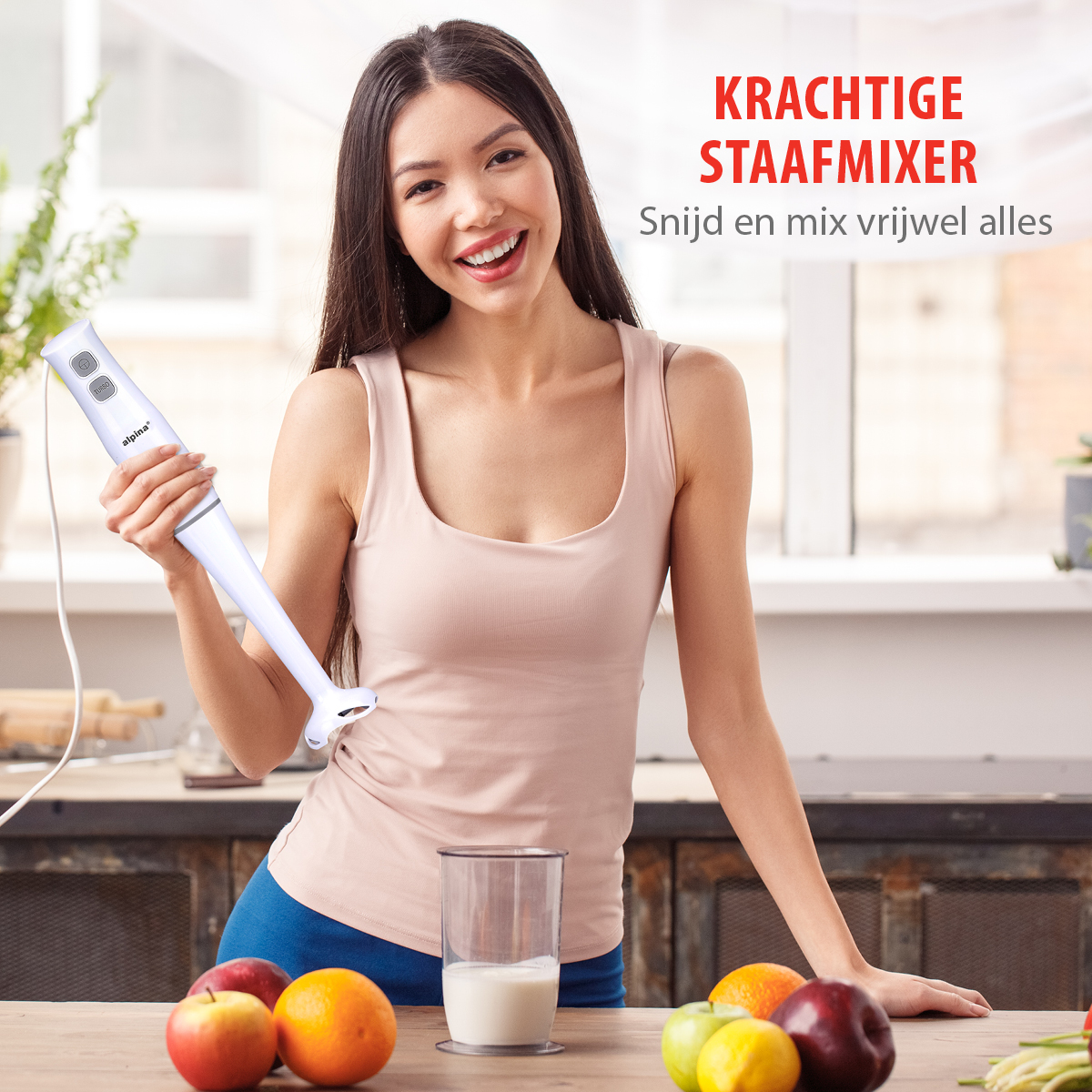 Vrouw met een witte staafmixer, lachend, met fruit en een glas melk op het aanrecht.